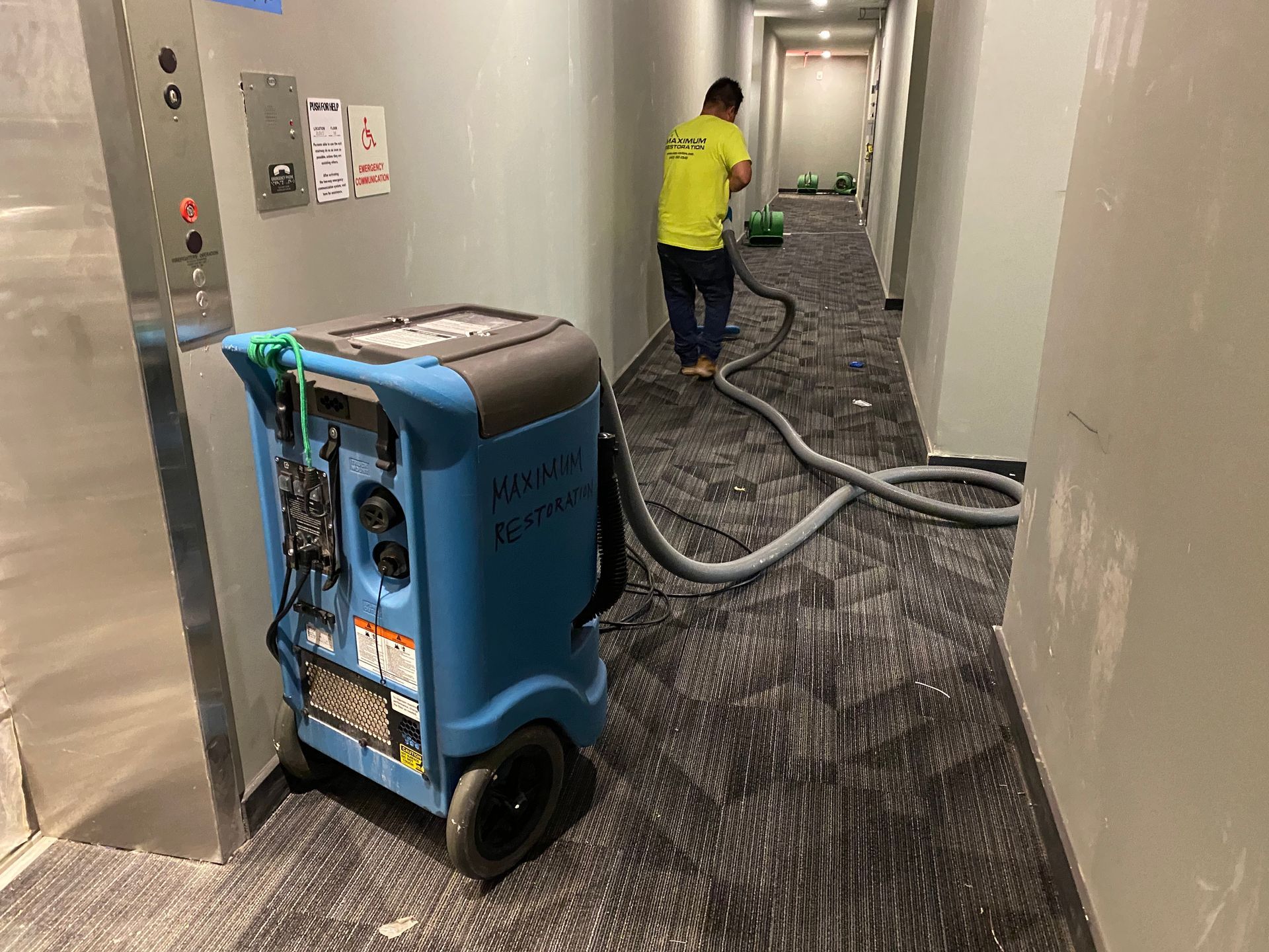 A worker in a neon yellow shirt uses a carpet cleaning machine in a hallway with dark patterned carpeting.