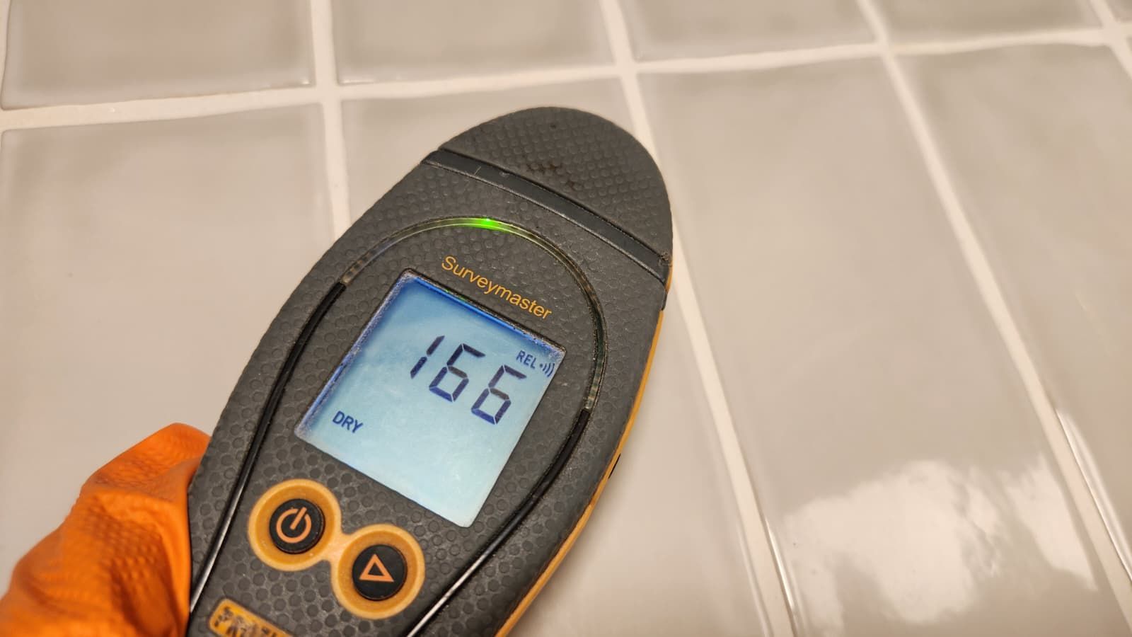 A hand in an orange glove holds a digital moisture meter against a tiled wall, displaying a reading of 166.