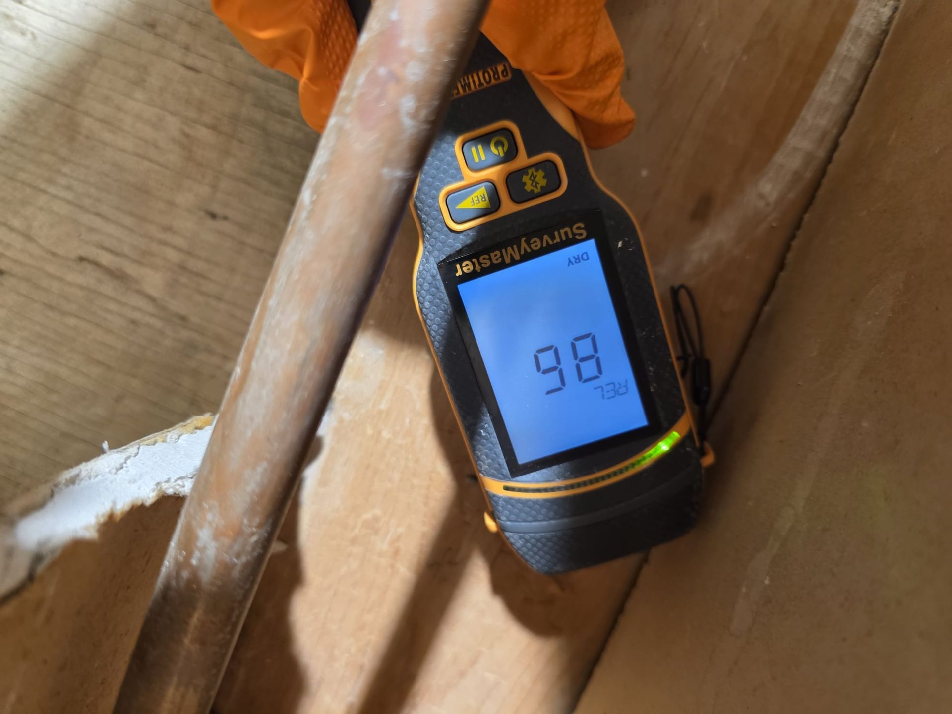 A moisture meter held against a wooden surface, displaying a reading of 98 in a brightly lit, dimly enclosed space.