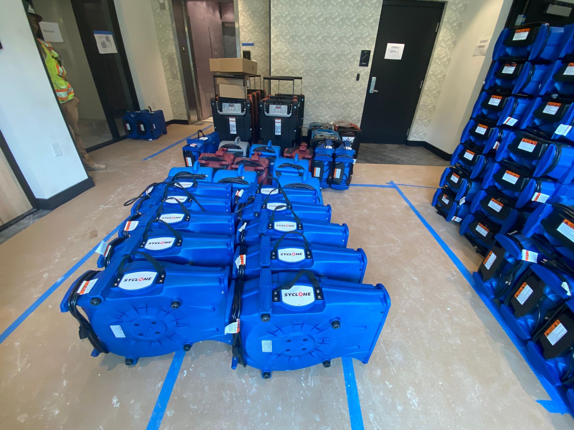 Rows of blue industrial air movers set up on a floor inside a room, with additional units stacked against a wall.