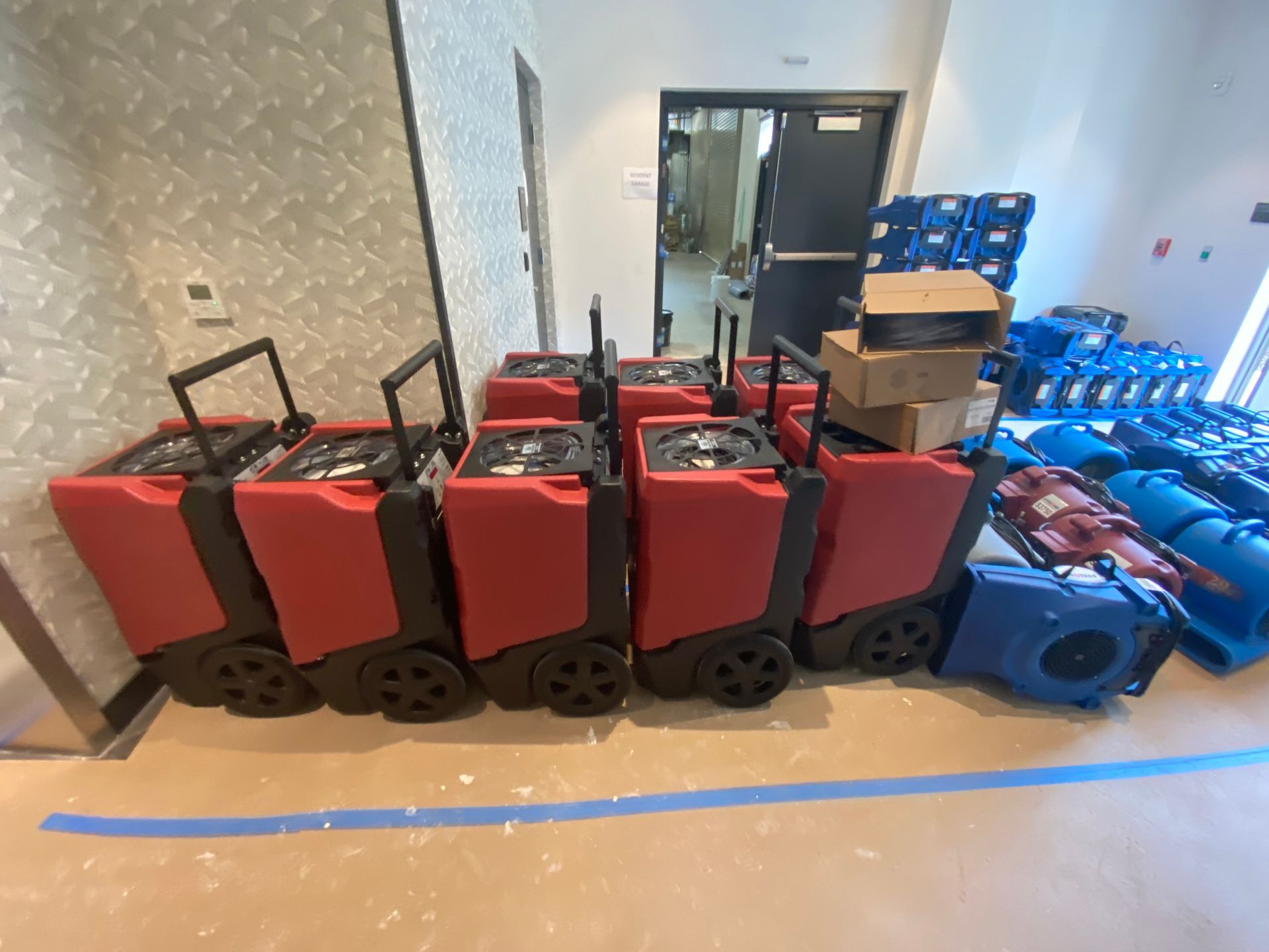 A row of red industrial dehumidifiers and a stack of blue floor fans in a room for water damage restoration.