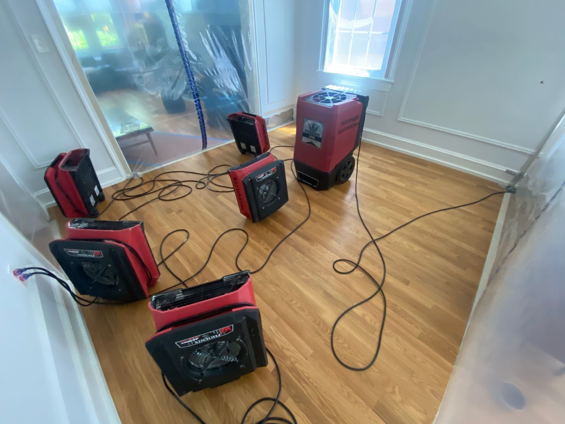 Red professional drying fans and a dehumidifier are placed on a hardwood floor in a room undergoing water damage repair.