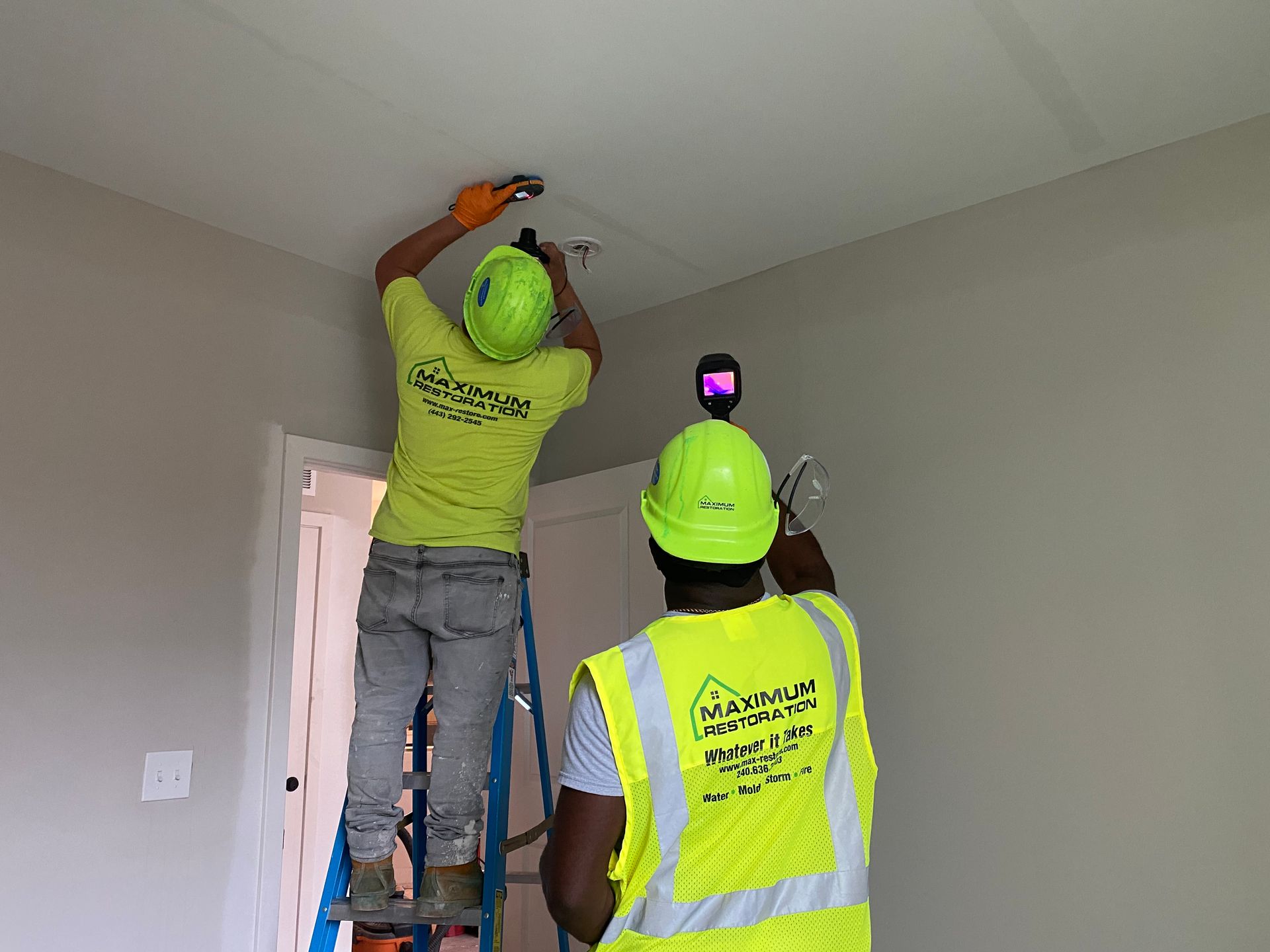 Two workers in high-visibility gear on a ladder inspecting a ceiling fixture with a thermal imaging device.