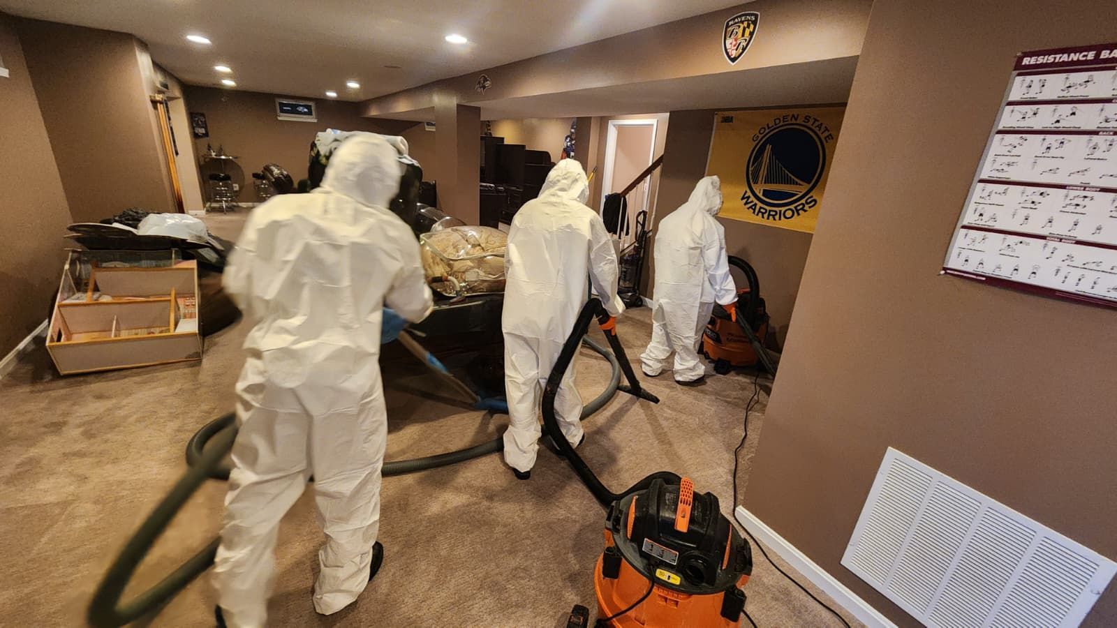 Three people in white hazmat suits vacuum the carpet in a finished basement with a Golden State Warriors flag on the wall.