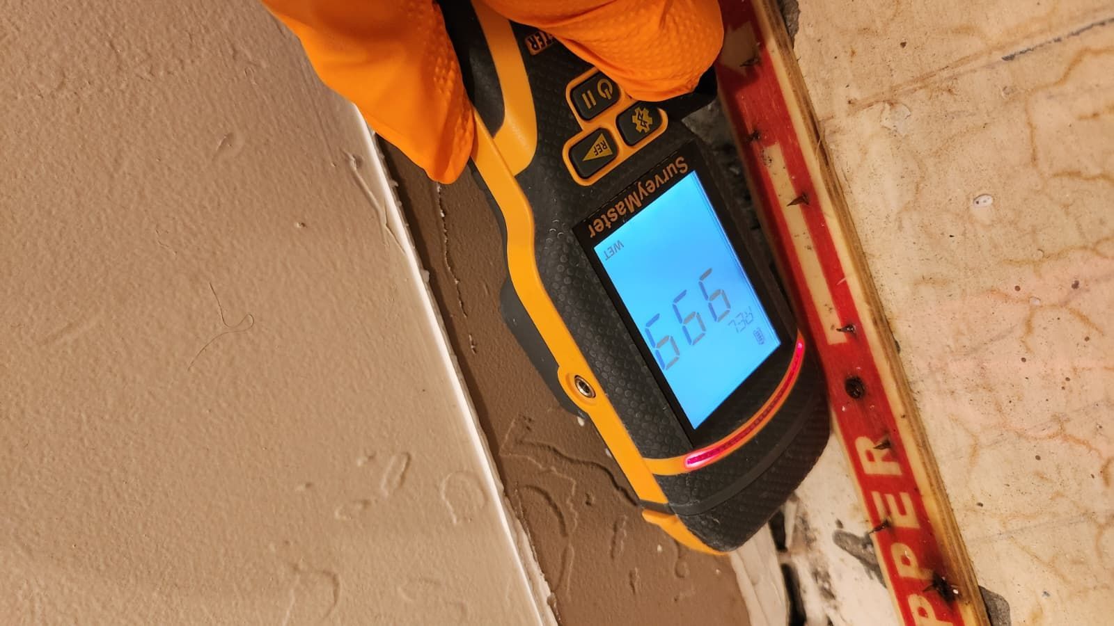 A hand wearing an orange glove holds a digital moisture meter against a wall, showing a reading of 99.9.