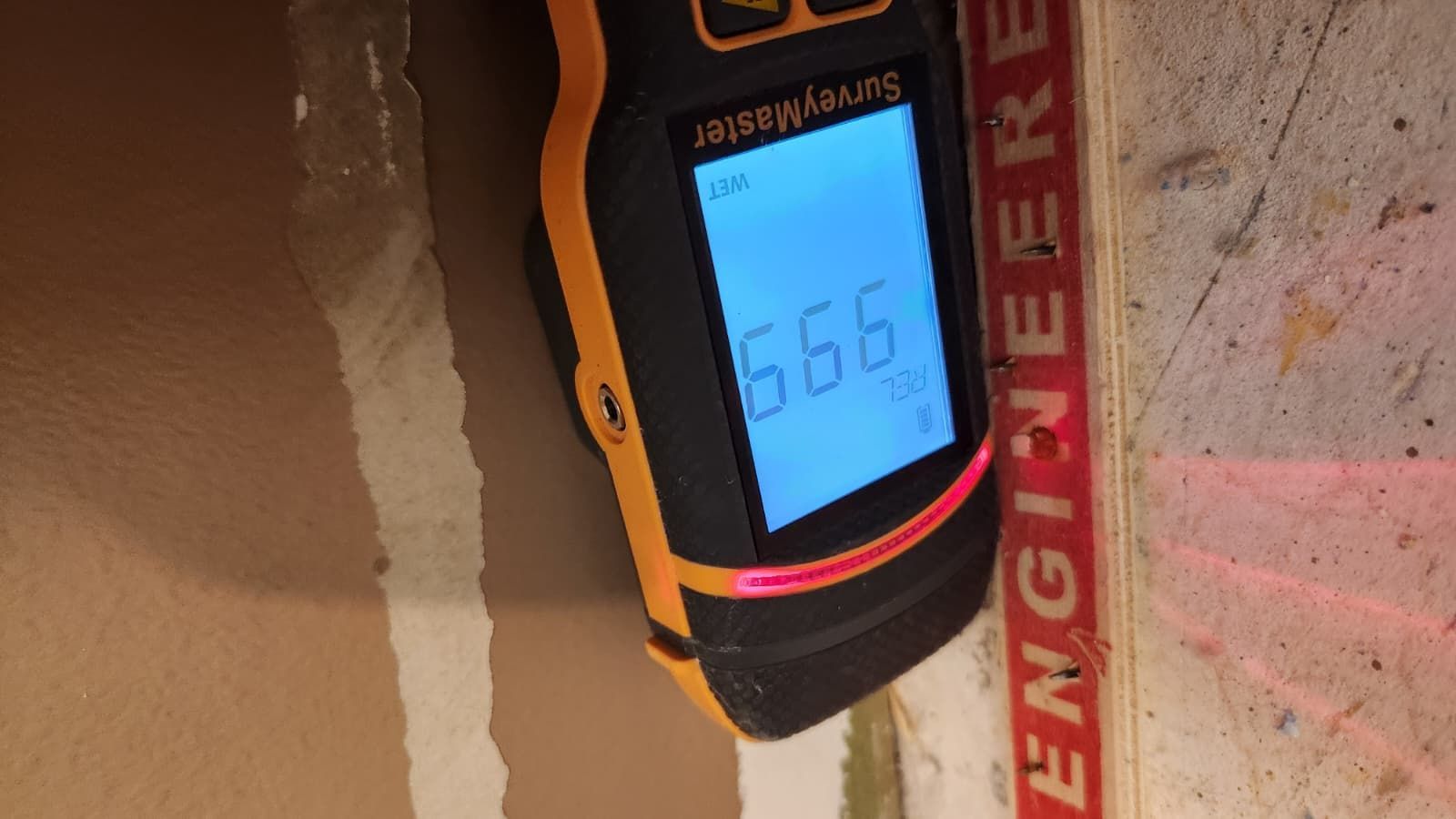 A yellow moisture meter held against a wooden wall shows a digital reading of 99.9.