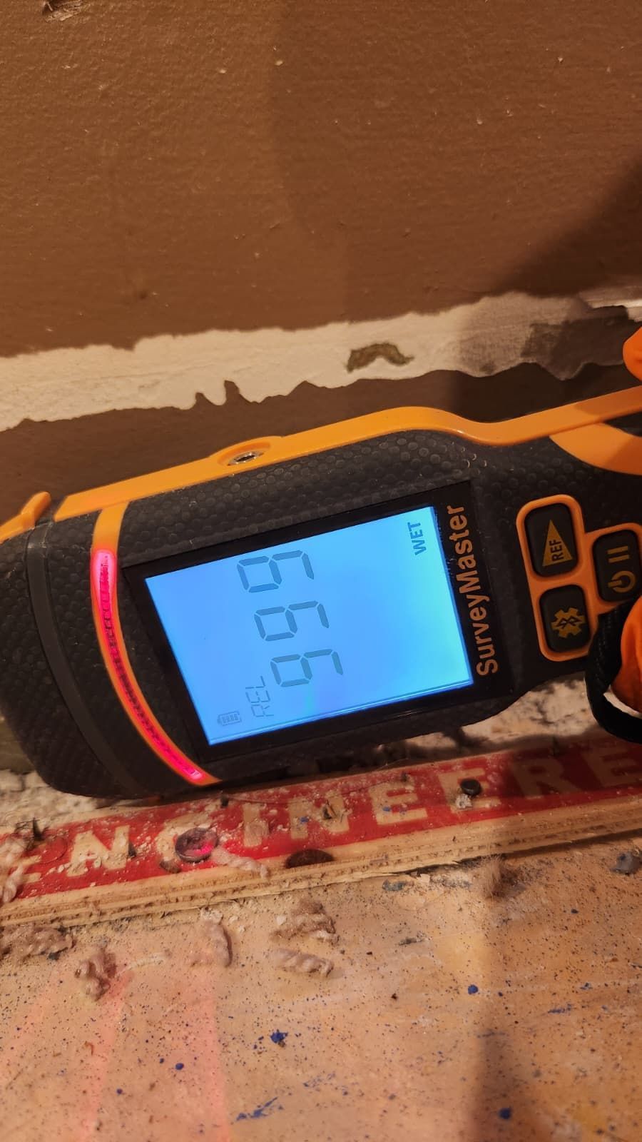 A yellow and black moisture meter displays the reading 99.9 on its digital screen while resting on a wood subfloor.