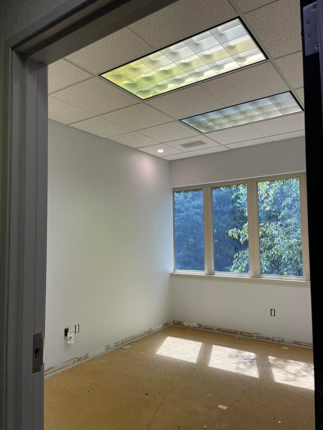 Empty office room with white walls, tiled ceiling lights, windows, and a bare floor.