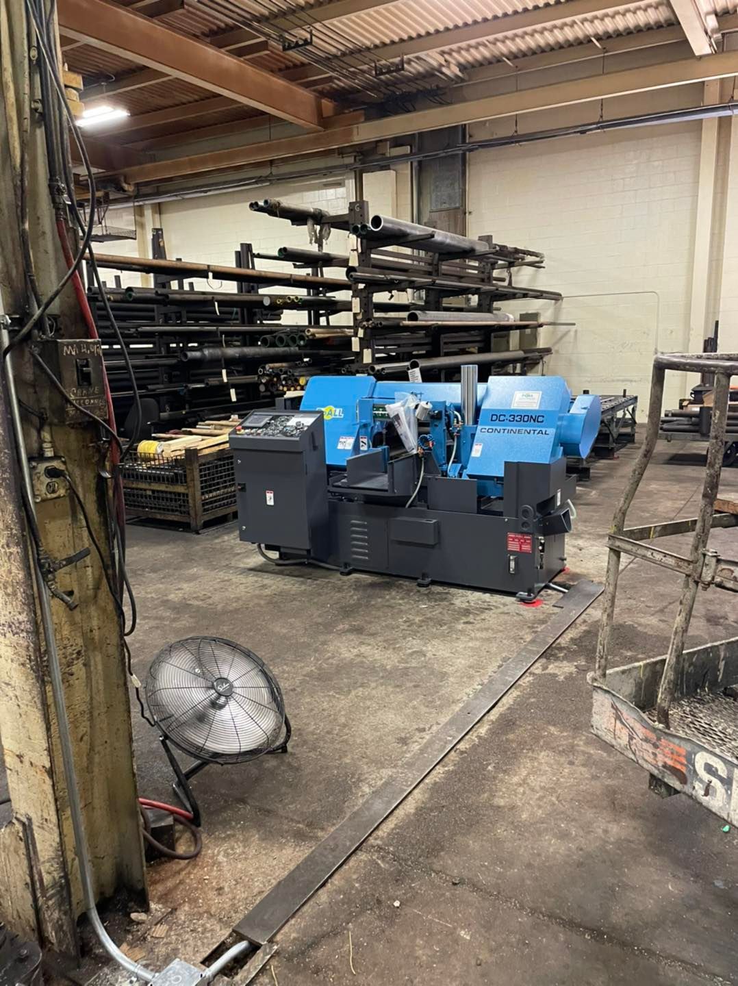 Industrial workshop with blue machinery, metal racks, and a floor fan under a corrugated roof