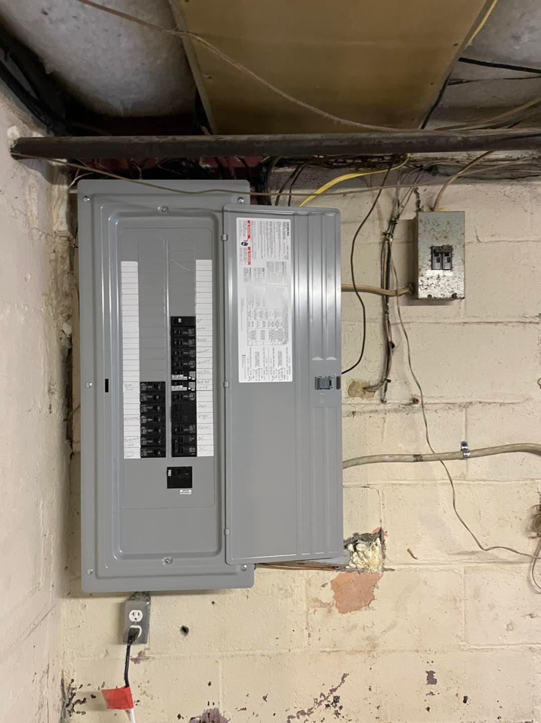 Gray electrical breaker panel on a basement wall beneath exposed pipes and wiring.