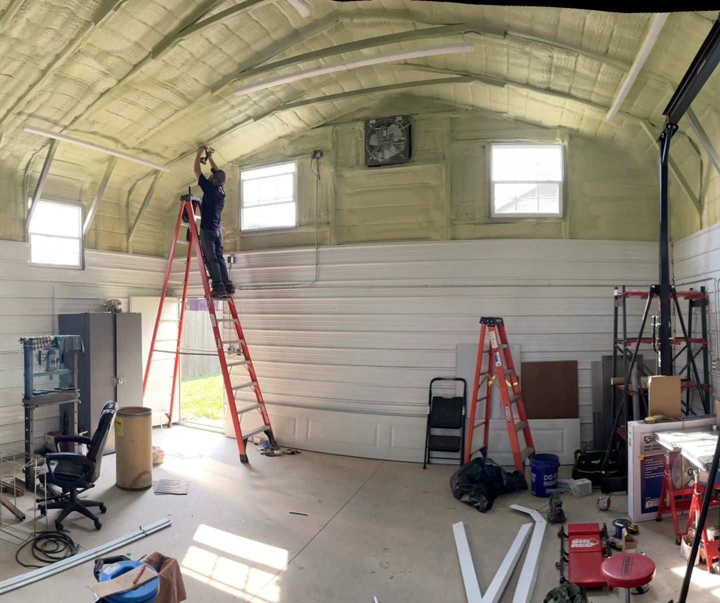 Person on a red ladder in a small workshop under a curved ceiling, with tools and equipment around.