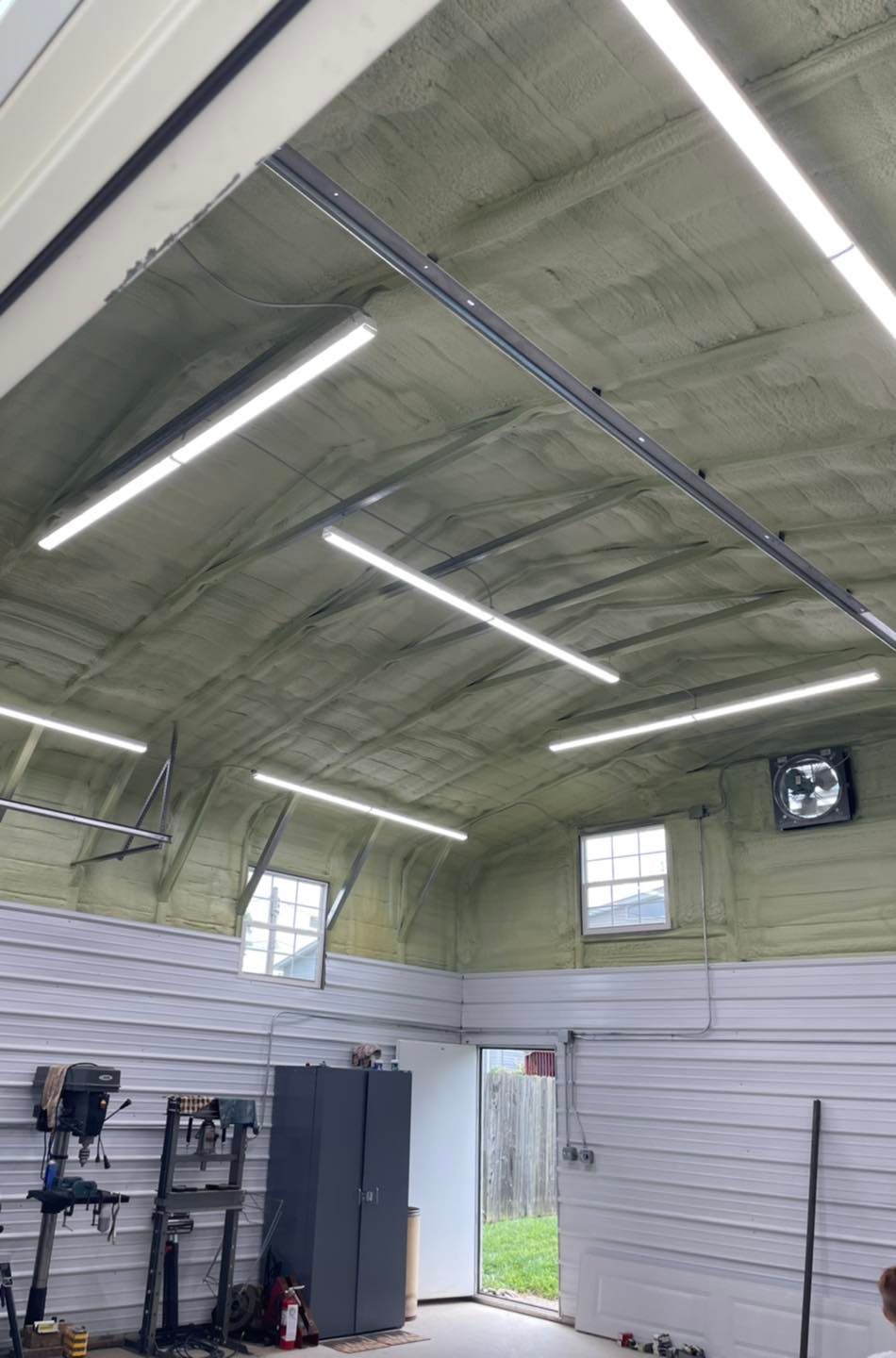 Interior of a workshop with a high arched ceiling, fluorescent lights, shelves, cabinets, and an open door.
