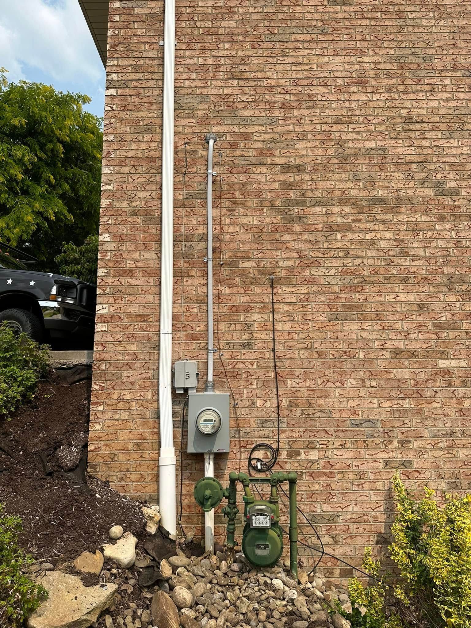 Brick exterior wall with utility meters, pipes, and bushes beside a parked vehicle