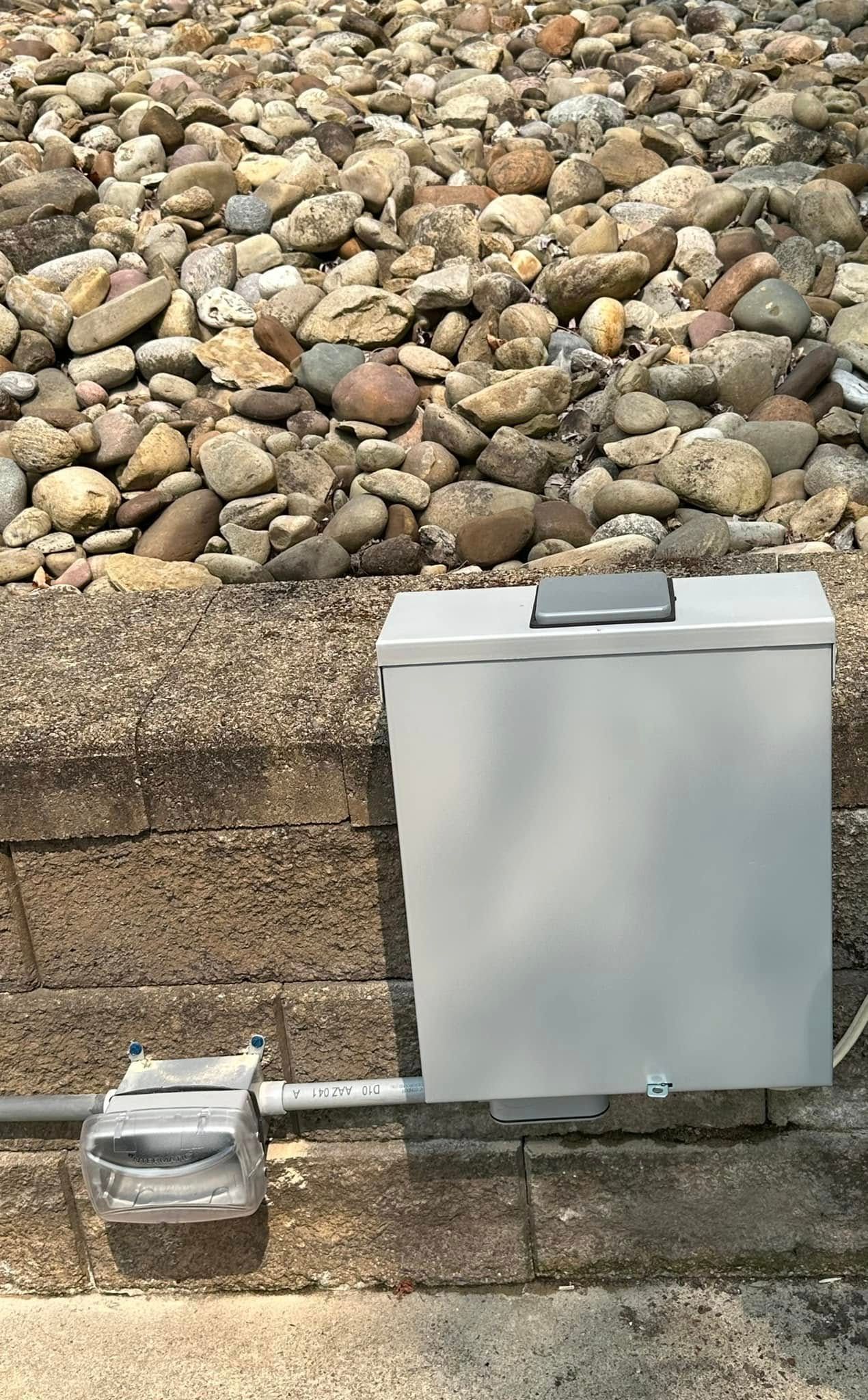 Gray electrical box on a stone wall with conduit, against a background of small rocks
