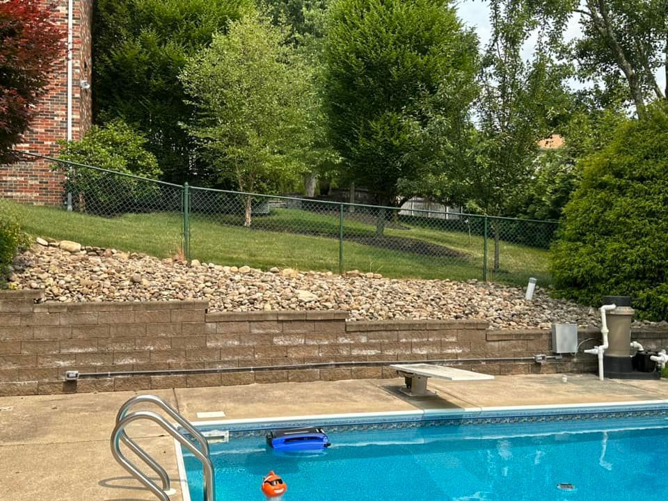 Backyard swimming pool with stone wall, deck chairs, and lush trees behind a fence