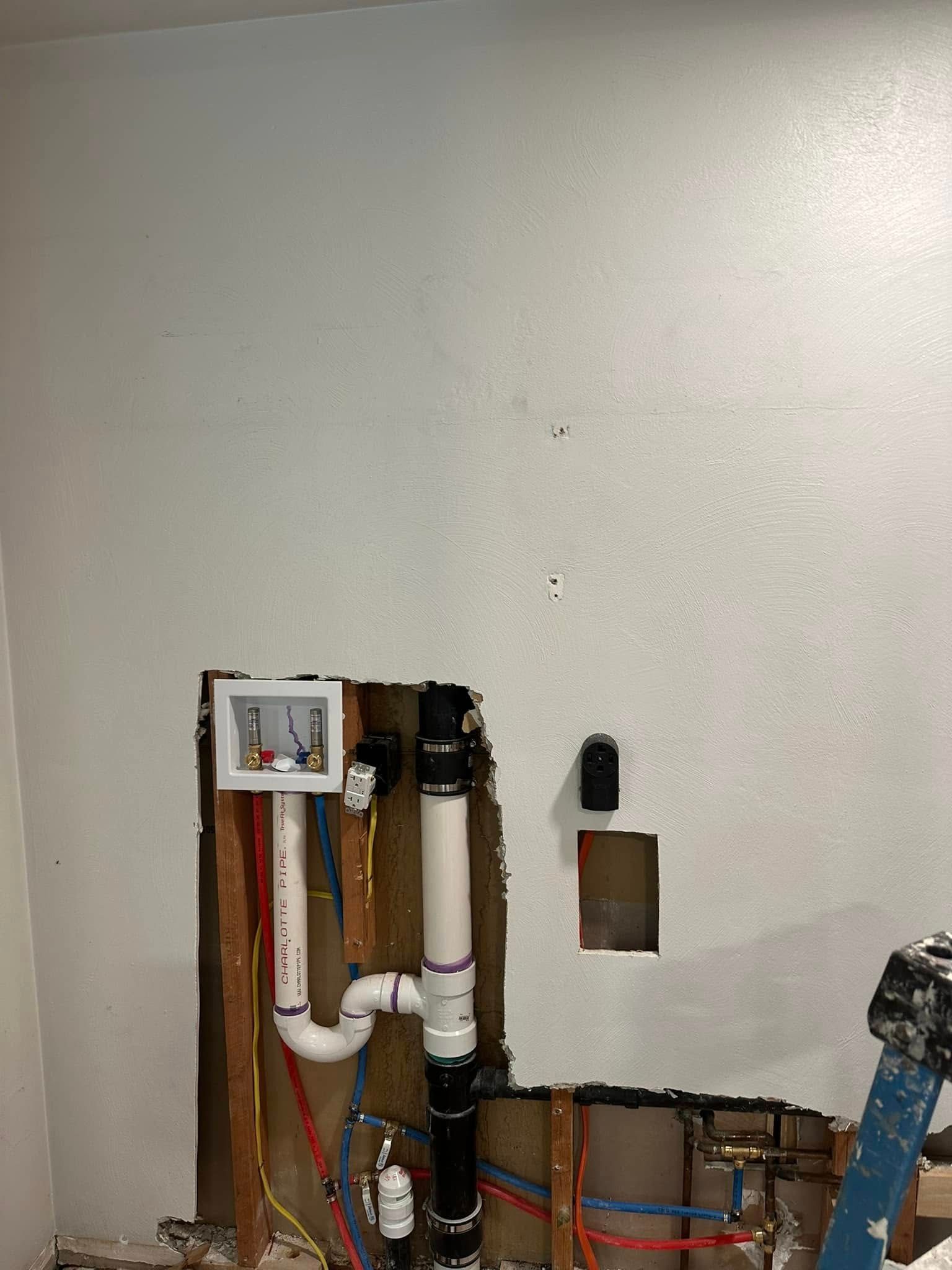Wall opening showing exposed plumbing pipes and fittings against a white wall, with a small utility cutout to the right