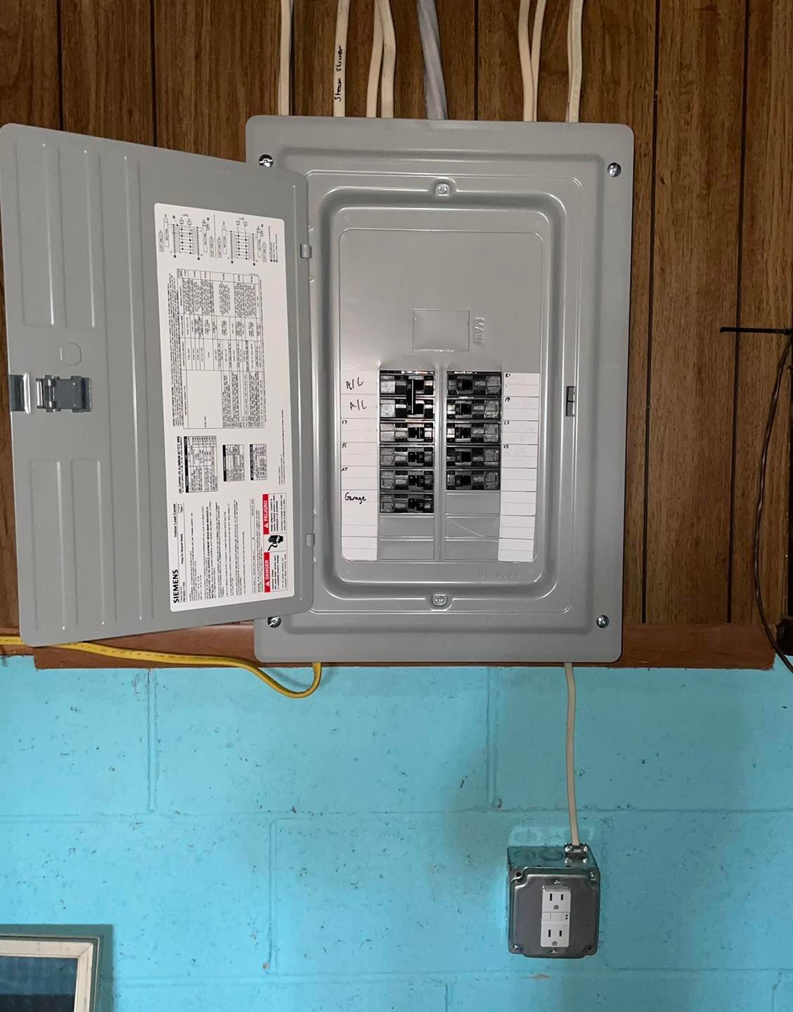 Open electrical breaker panel on a wood wall above a blue painted section, with a corded device below.