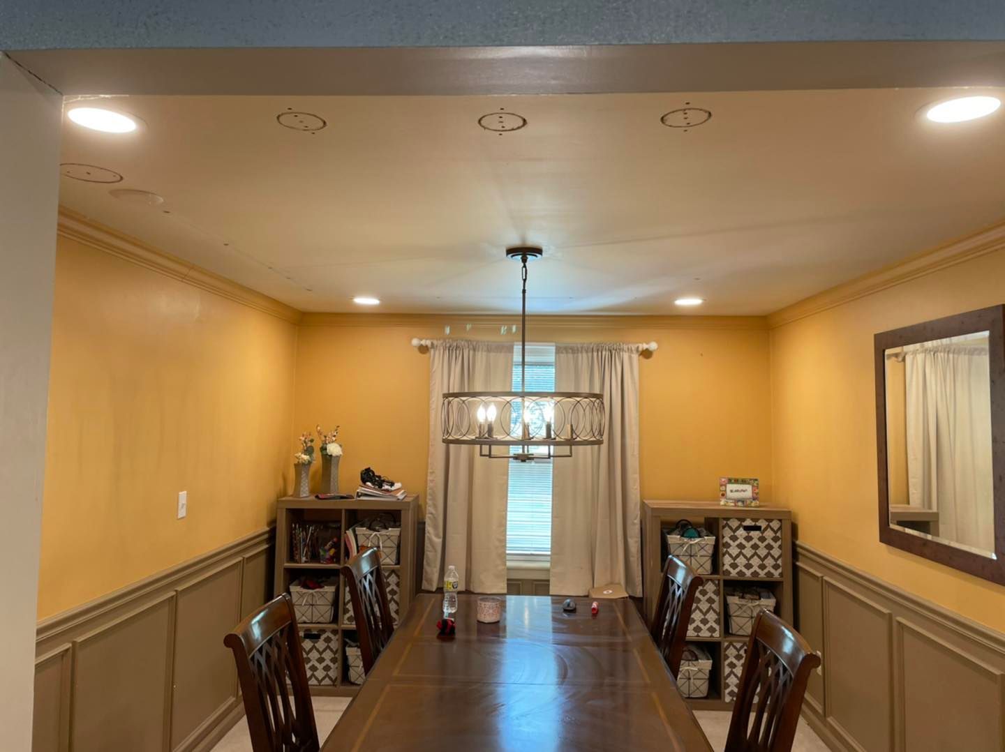 Dining room with a long wooden table, chairs, beige walls, chandelier, and window curtains.