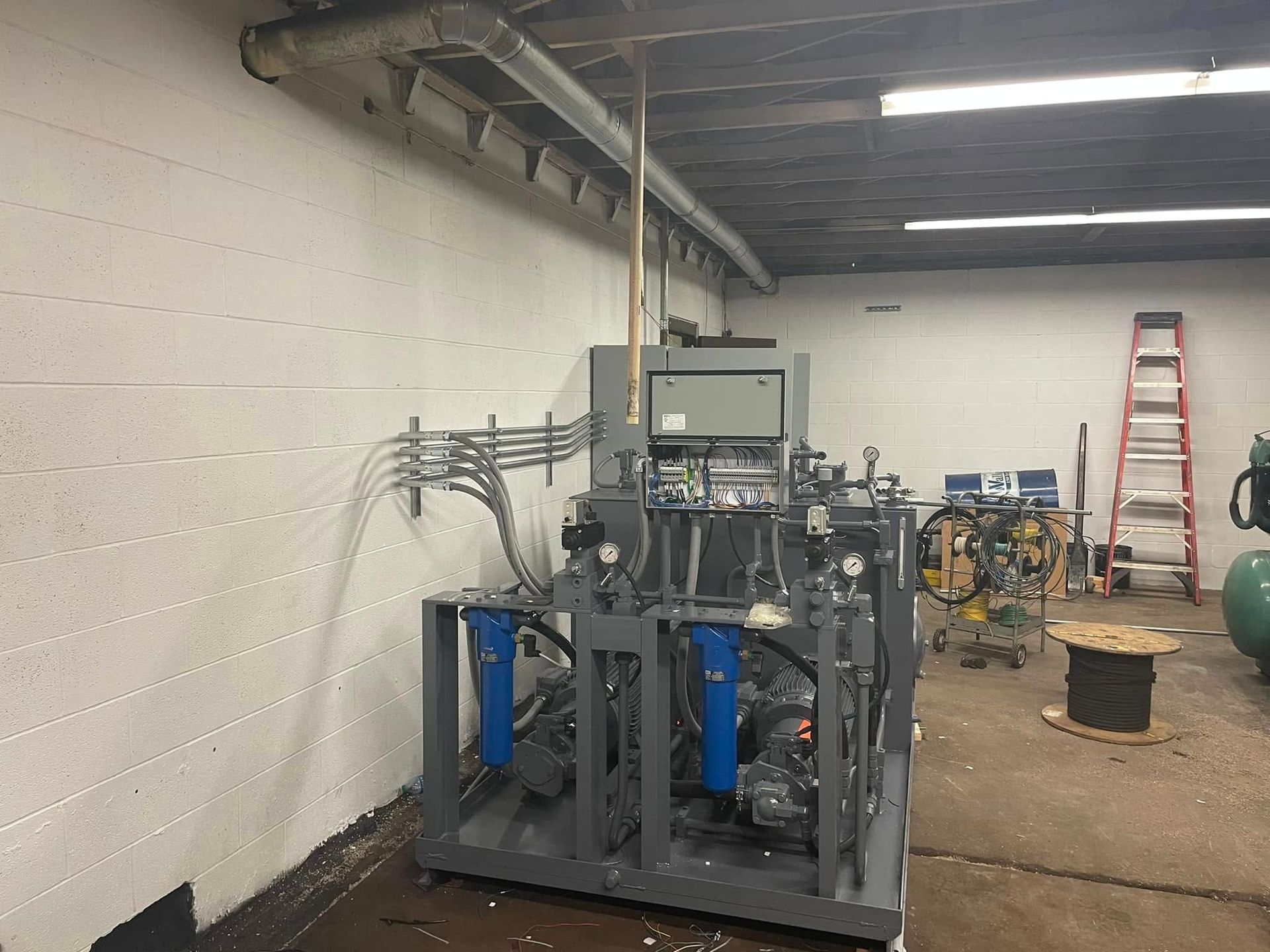 Industrial machine setup in a warehouse, with metal framing, hoses, and a red ladder in the background