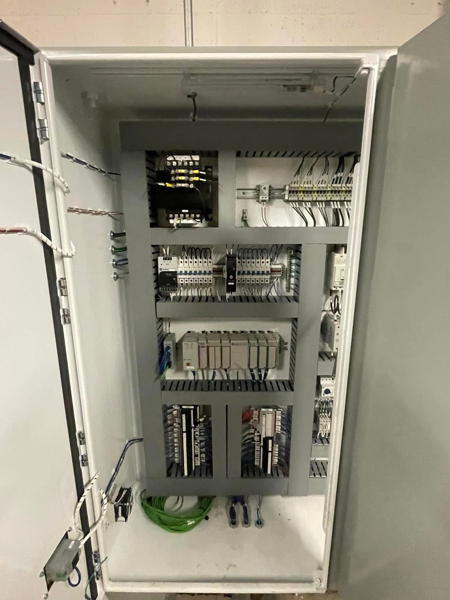 Open electrical control cabinet with wiring, relays, circuit breakers, and components on metal shelves.