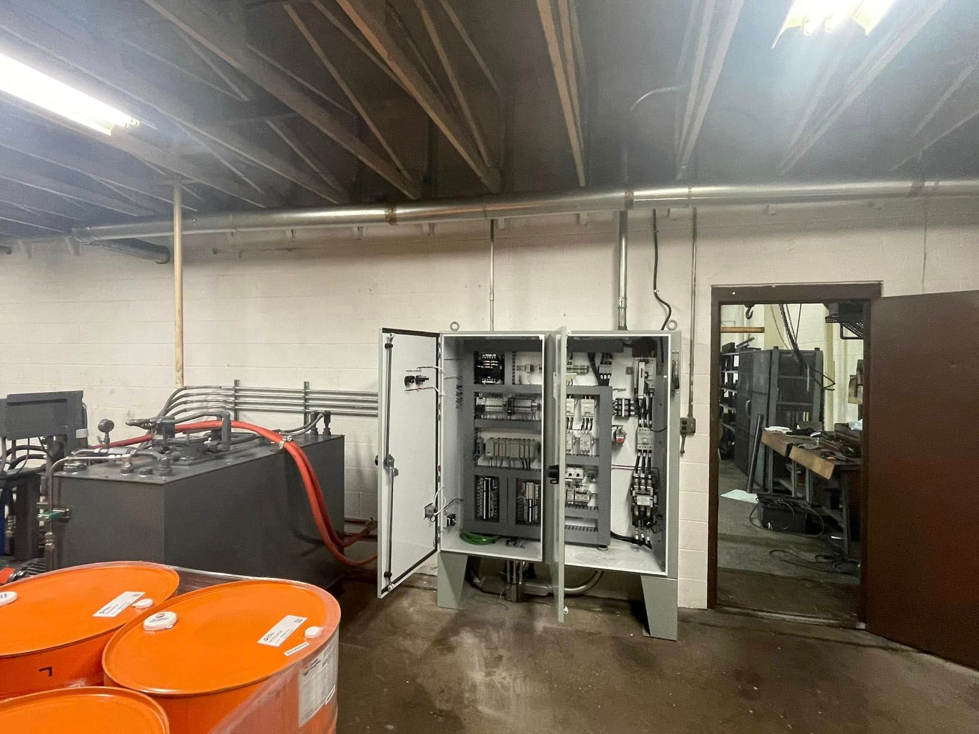 Industrial room with open electrical cabinet, orange barrels, and equipment under a metal ceiling