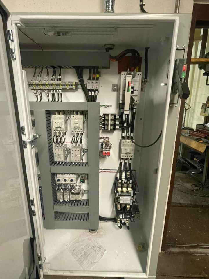 Open electrical control panel cabinet with wiring, relays, and circuit breakers inside a workshop.