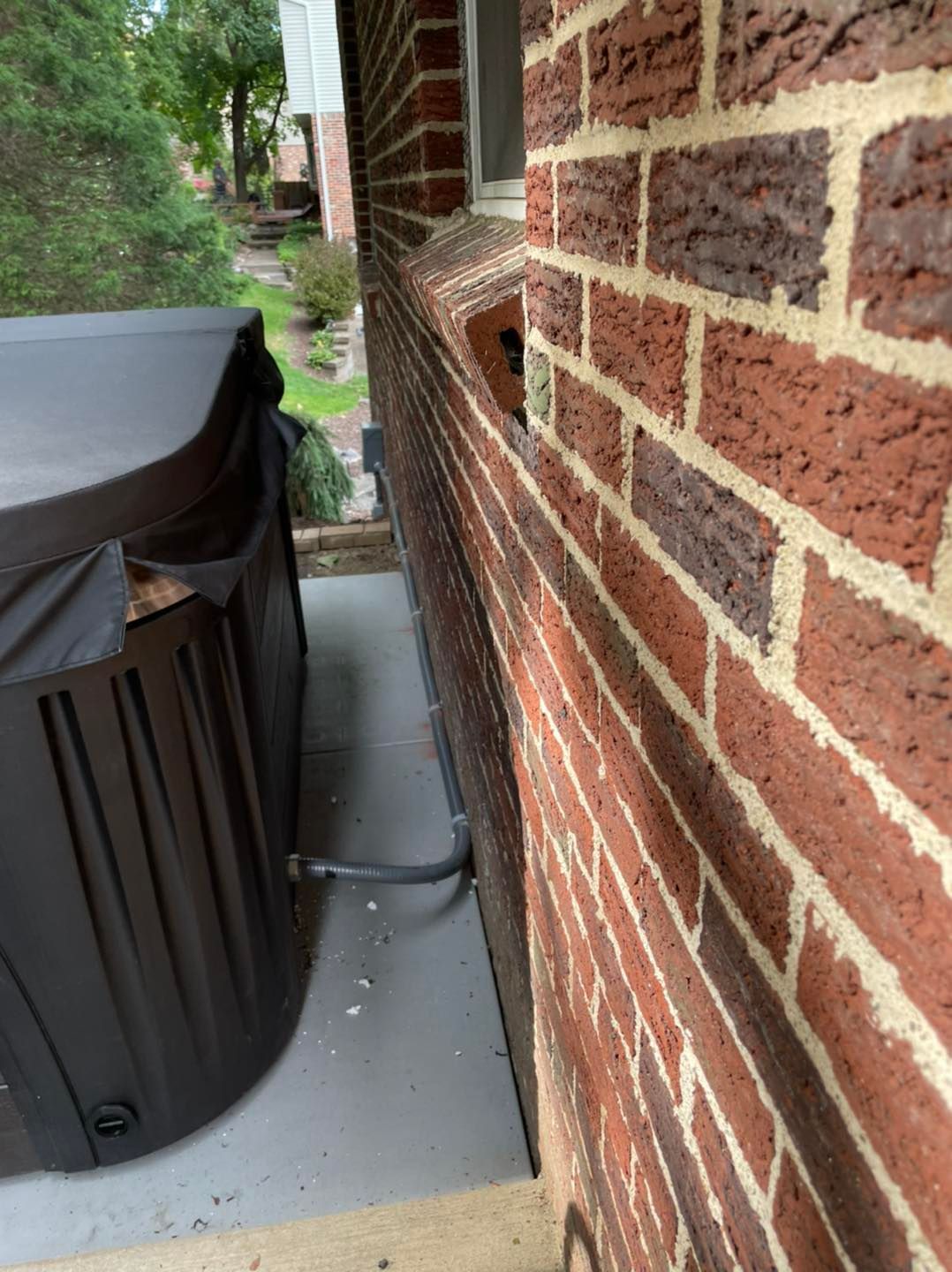 Brick house wall beside a covered patio with a large black trash bin on a concrete slab
