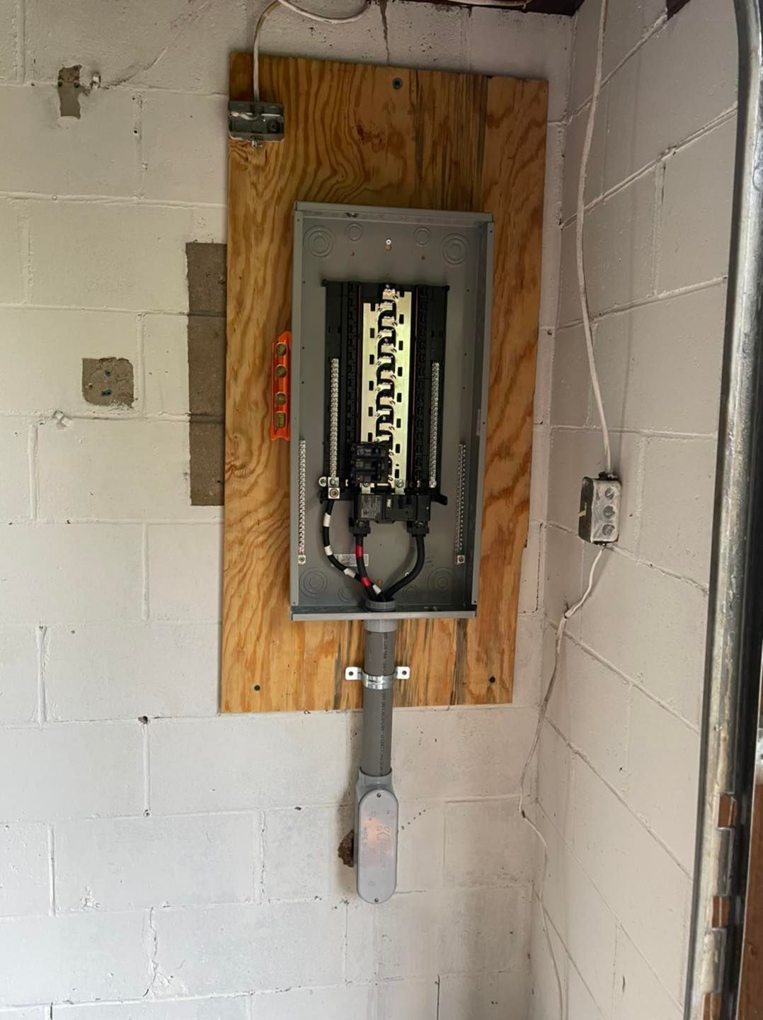 Electrical breaker panel mounted on plywood in a cinder block utility room.