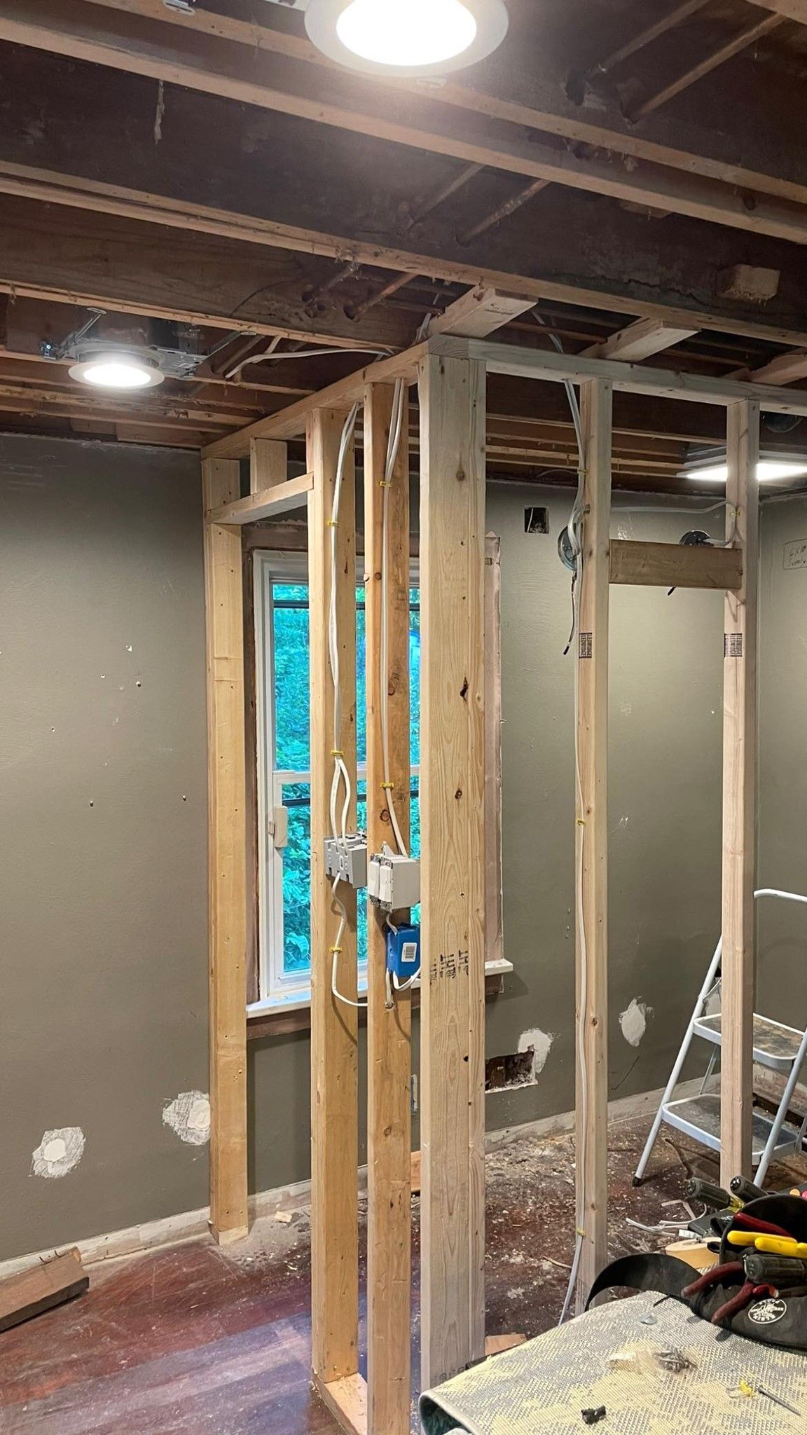 Partially framed interior room under renovation with exposed studs and drywall, ceiling lights, and tools on the floor