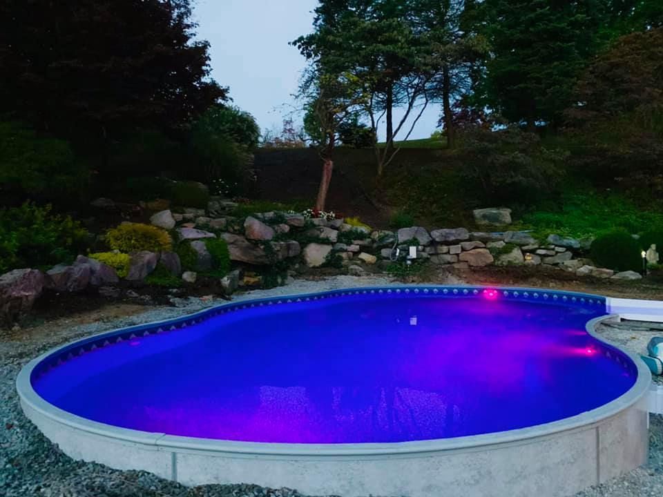 Backyard swimming pool glowing blue-purple at dusk, surrounded by rocks and trees