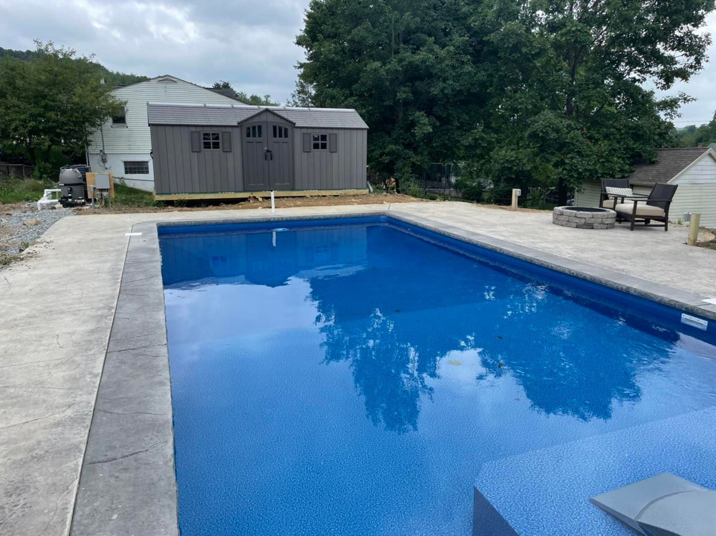 Blue backyard swimming pool beside a gray outbuilding and patio, with trees in the background