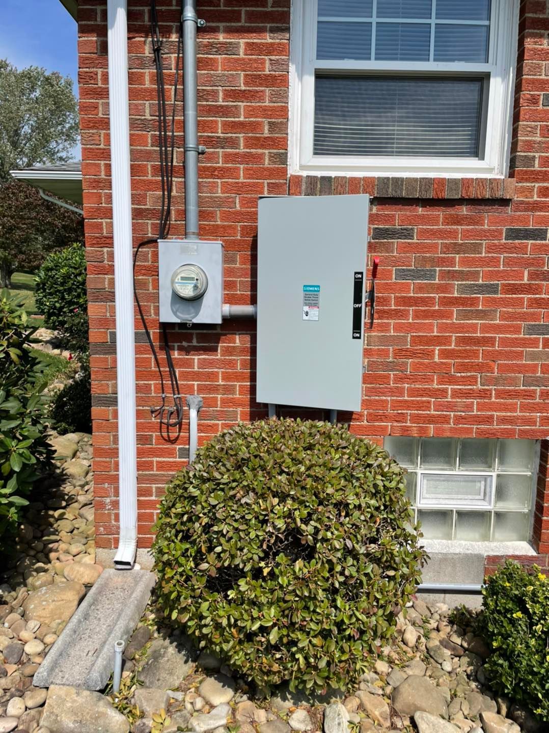 Brick house exterior with electric meter and gray utility panel beside a window and shrubs