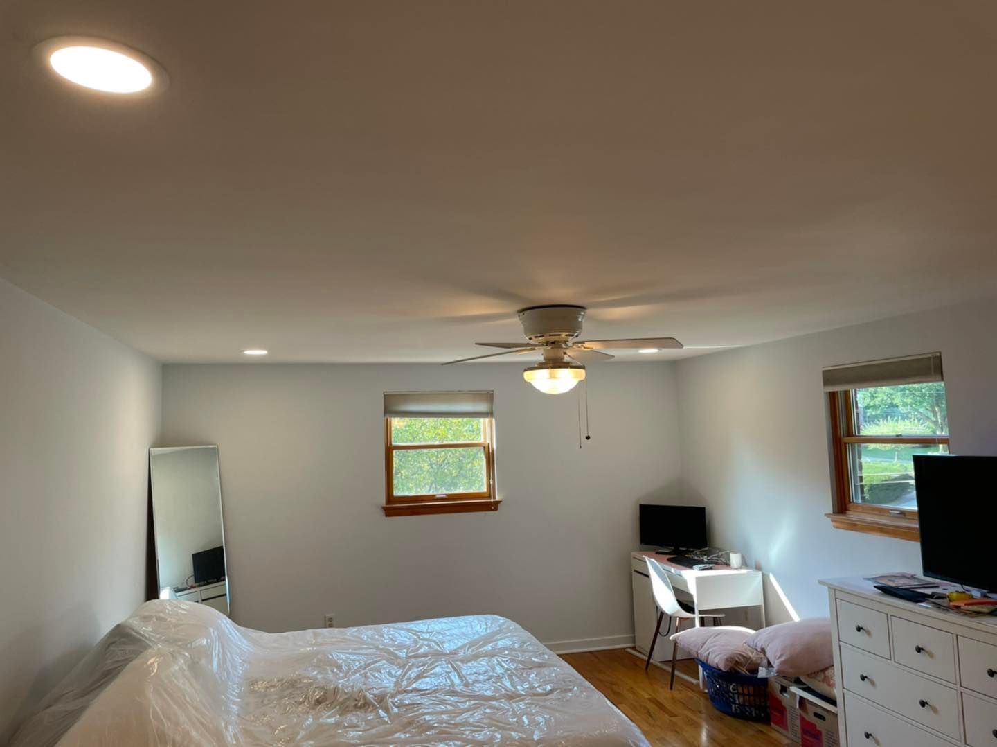 Bedroom with white walls, ceiling fan, bed, mirror, TV, and dresser in a bright room.