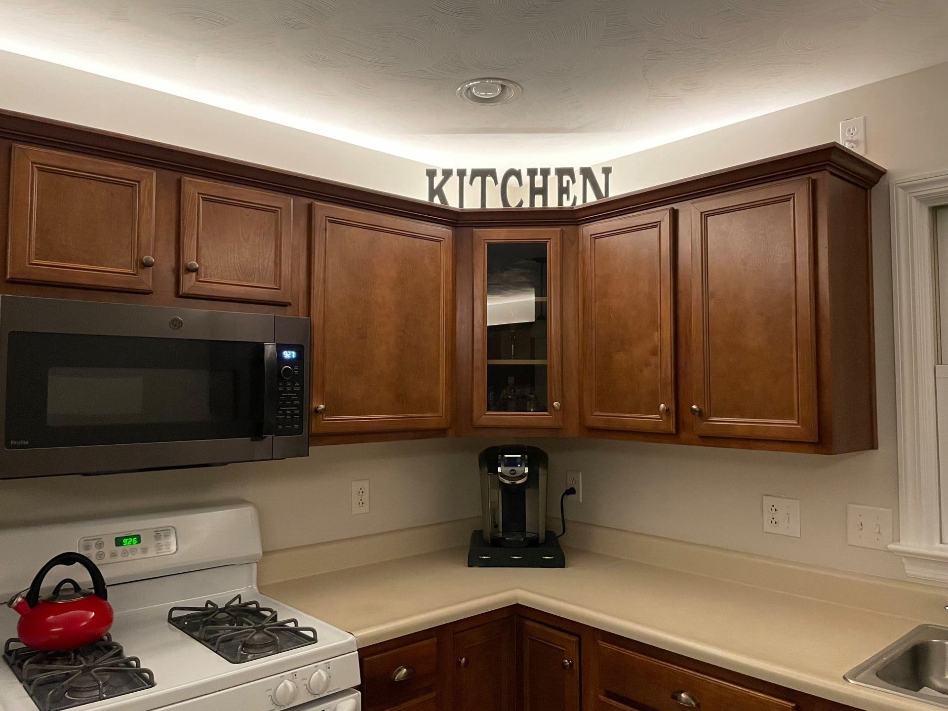 Compact kitchen with wooden cabinets, black microwave, stove, coffee maker, and a red kettle on the counter.