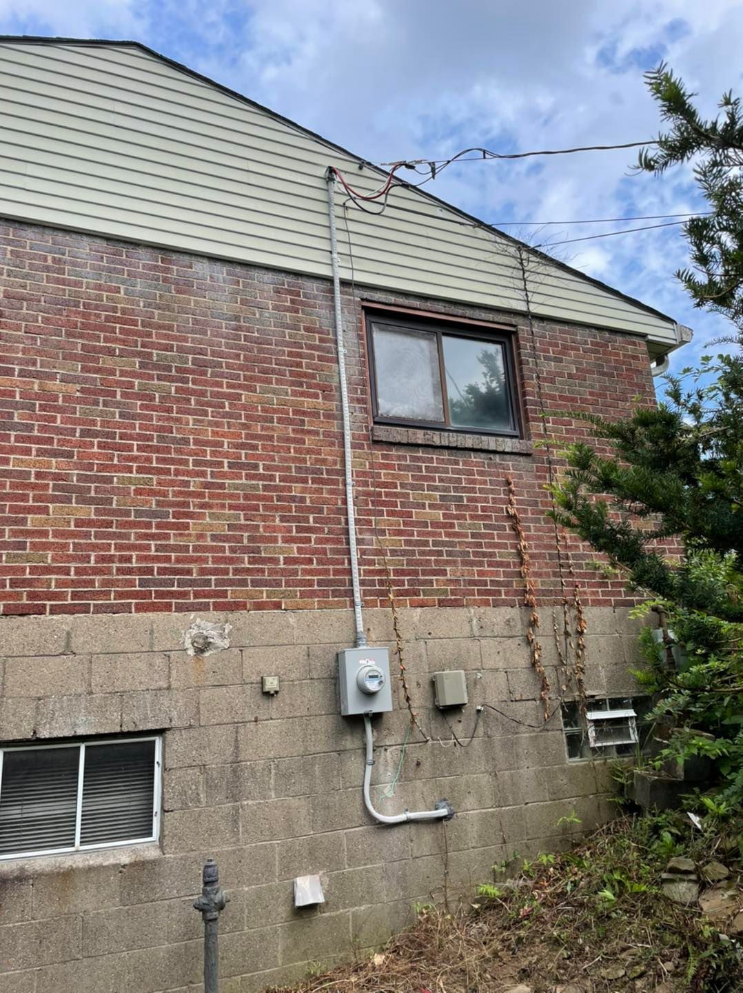 Brick and cinderblock house exterior with window, utility meters, and steep roof under cloudy sky