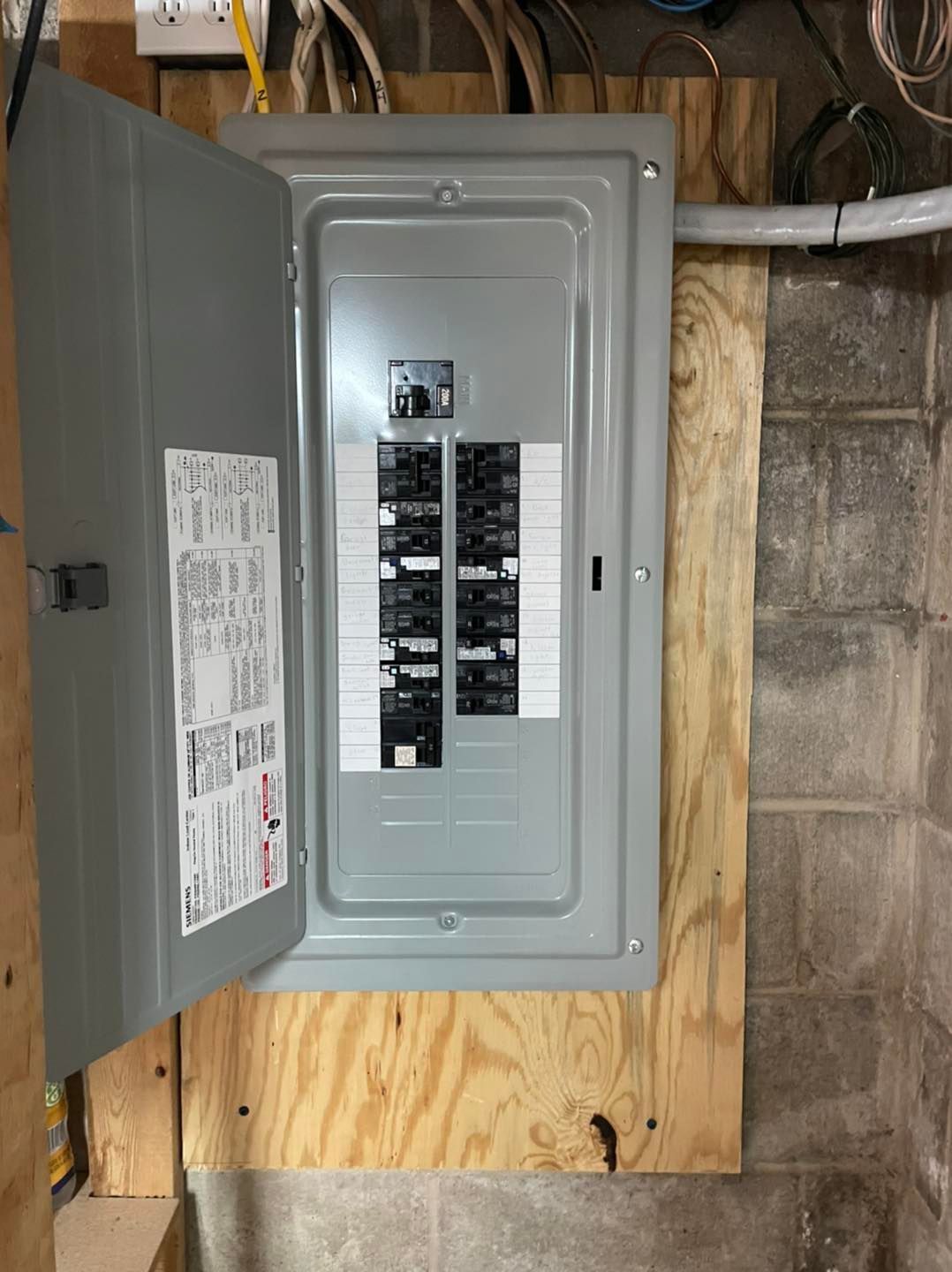 Open electrical breaker panel in a utility room with exposed wiring and plywood backing