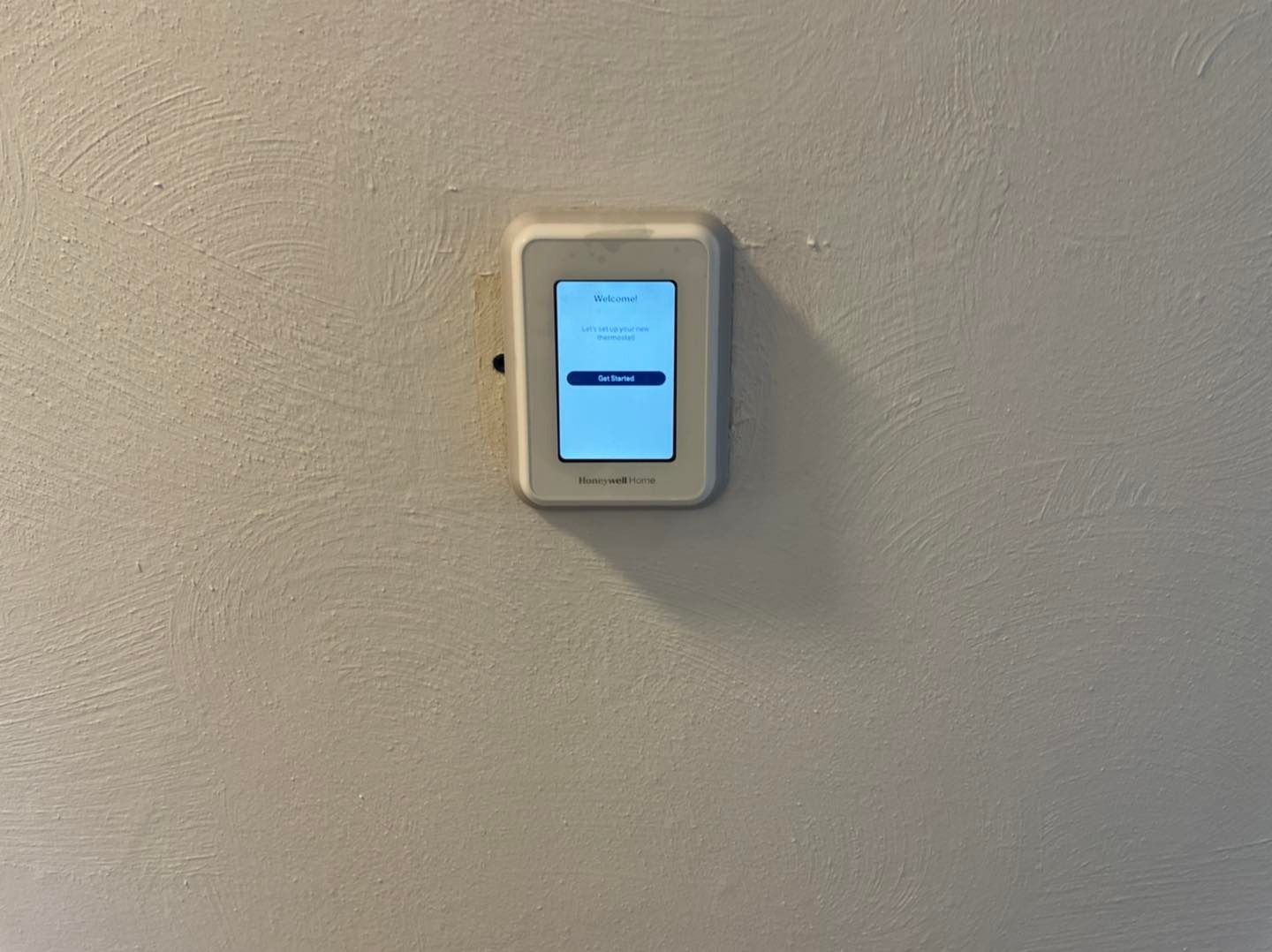 Wall-mounted touchscreen in a beige holder displaying a blue screen against a gray wall.