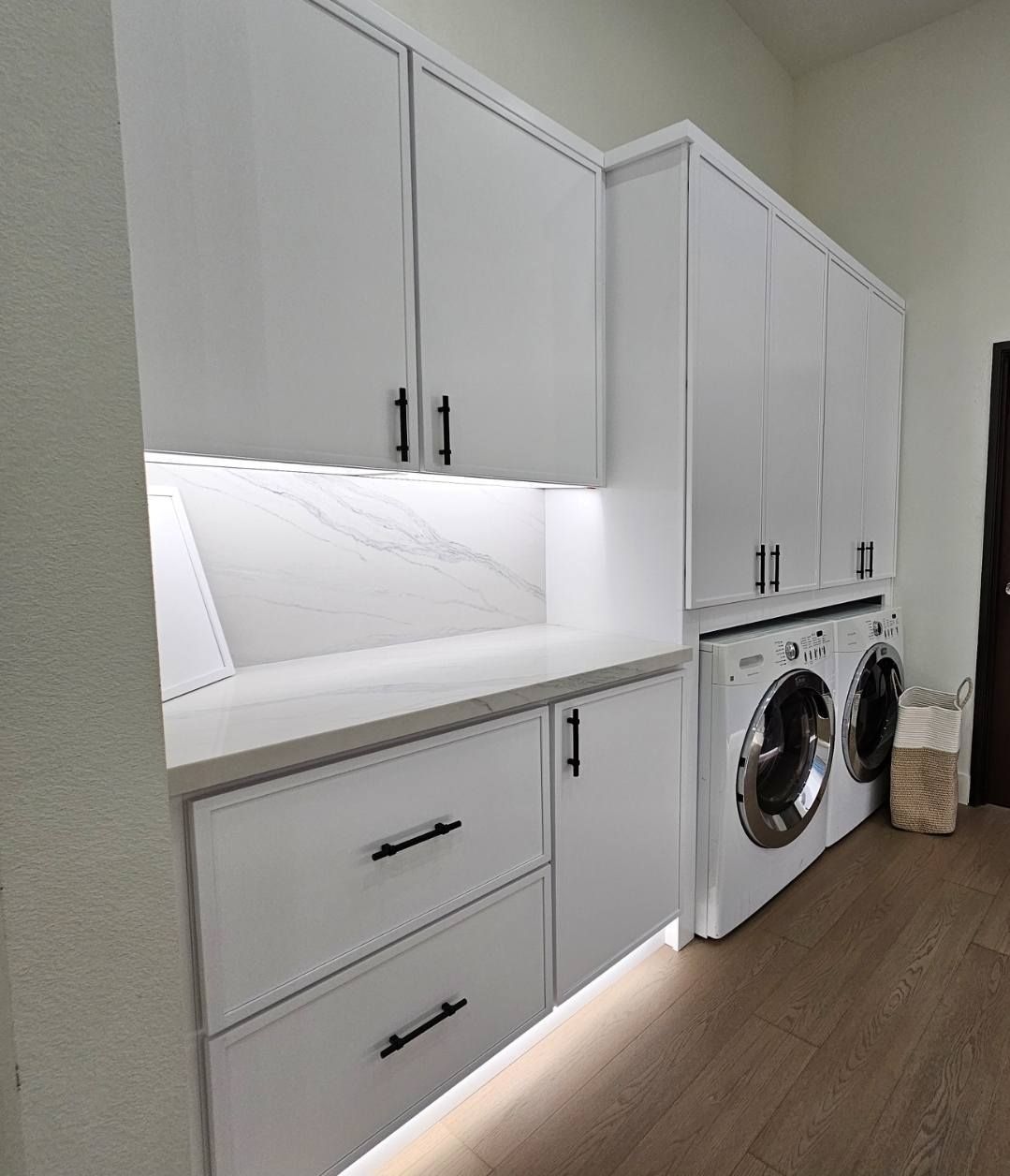 A white laundry room with cabinetry, a countertop, washer, and dryer. Cabinets have black hardware and a light strip beneath them.