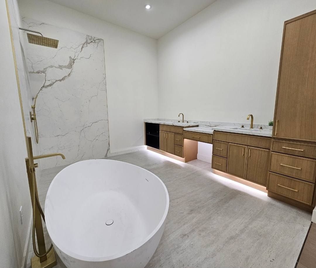 Modern bathroom featuring a white freestanding tub, marble shower, light wood cabinets with gold hardware, and light gray flooring.