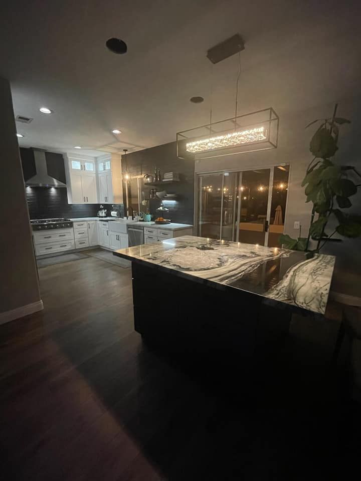 Dark kitchen with a large island, modern chandelier, and dark countertops. White cabinets and stainless steel appliances are visible in the background.