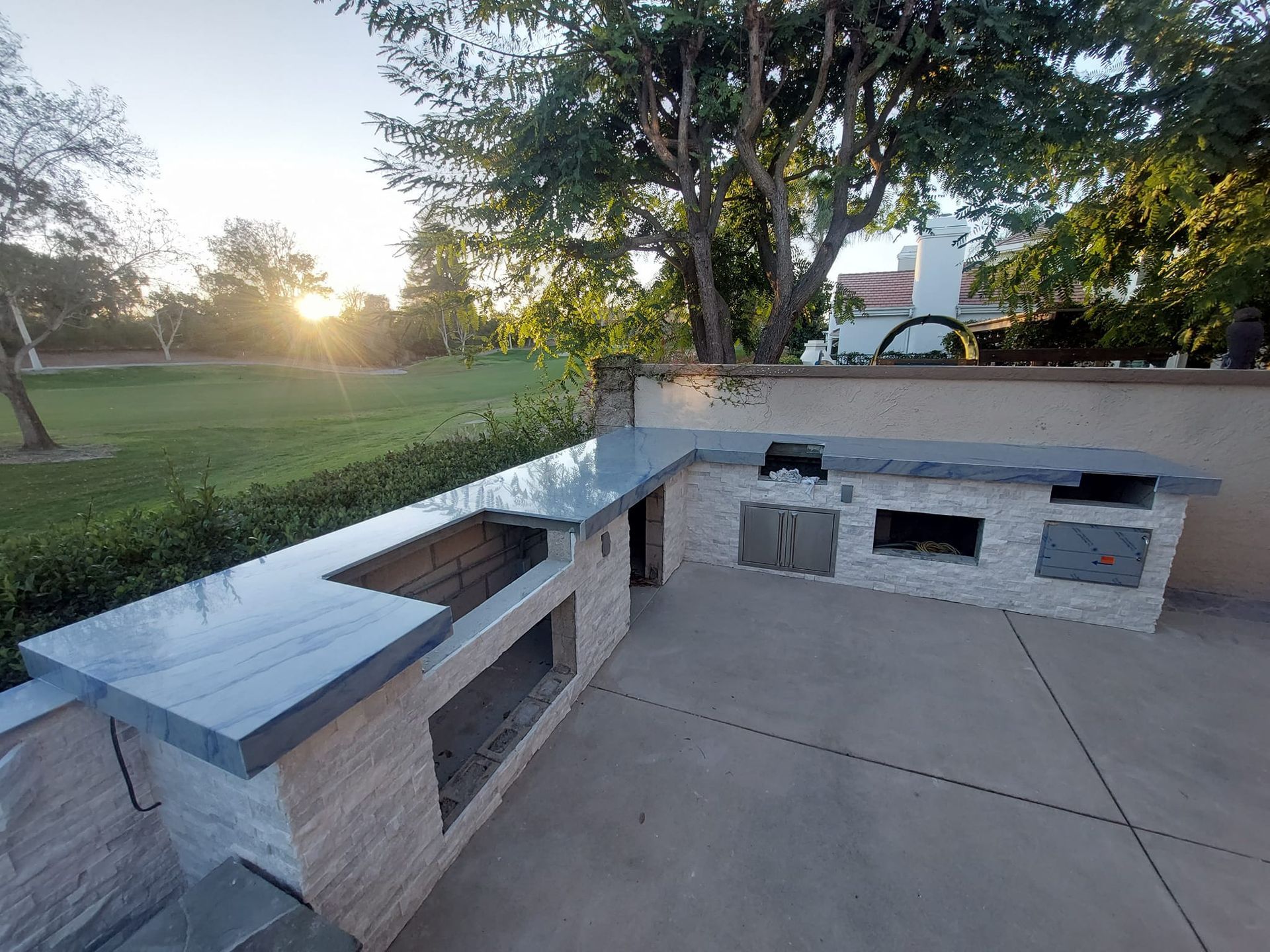 Outdoor kitchen with a gray countertop, built-in grill, and stainless steel appliances. Located on a patio, with trees and a grassy area in the background.