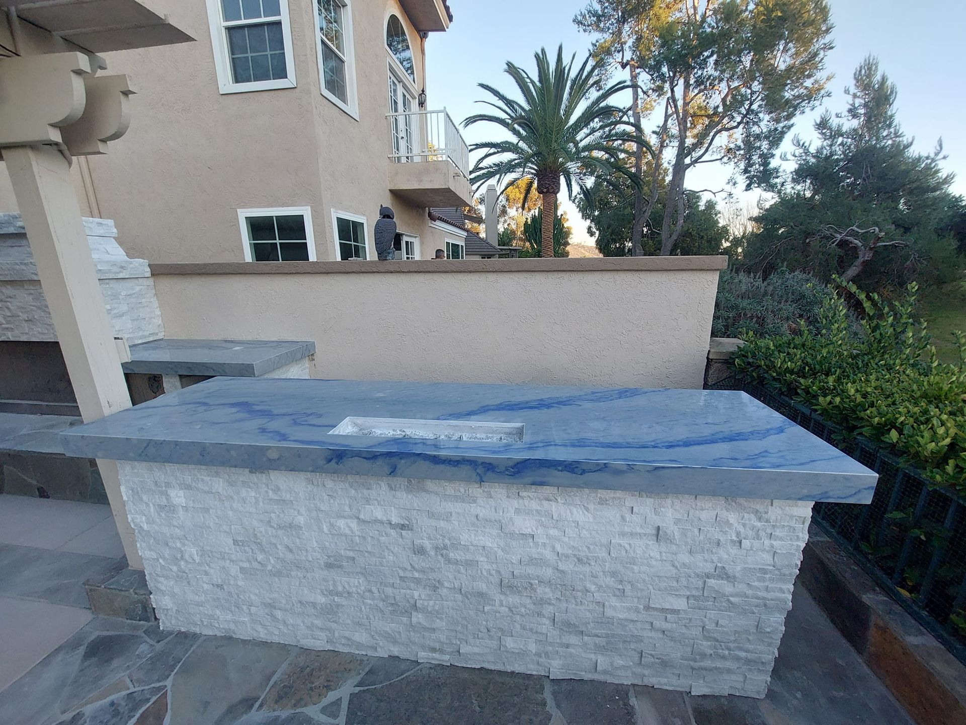 Outdoor stone fire pit with a blue granite top. The pit is in front of a two-story house with a palm tree in the background.