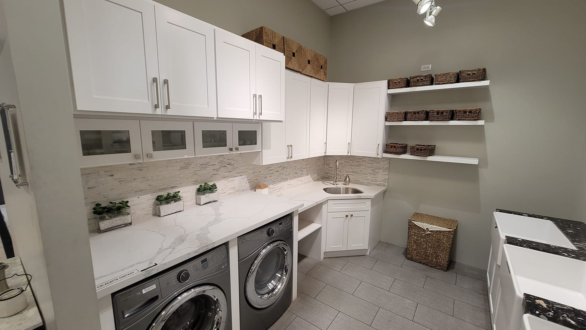 A well-lit laundry room with white cabinets, appliances, and a counter. There's a washing machine, a dryer, and shelves on the wall.