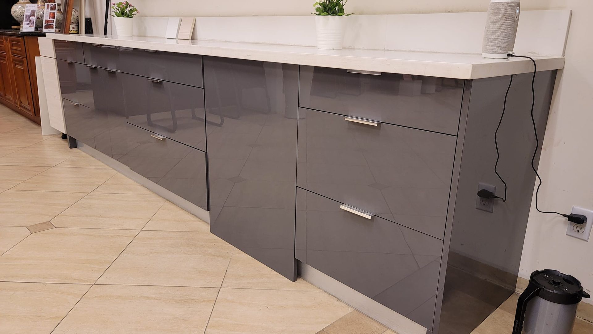 Long, modern kitchen cabinets in glossy gray with a white countertop. Two small plants and a paper towel roll sit on top.