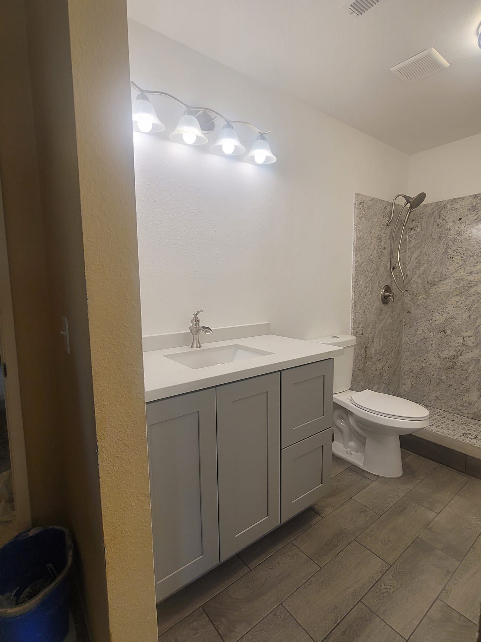 A bathroom with a gray vanity, toilet, and tiled shower. Overhead, a light fixture illuminates the space.