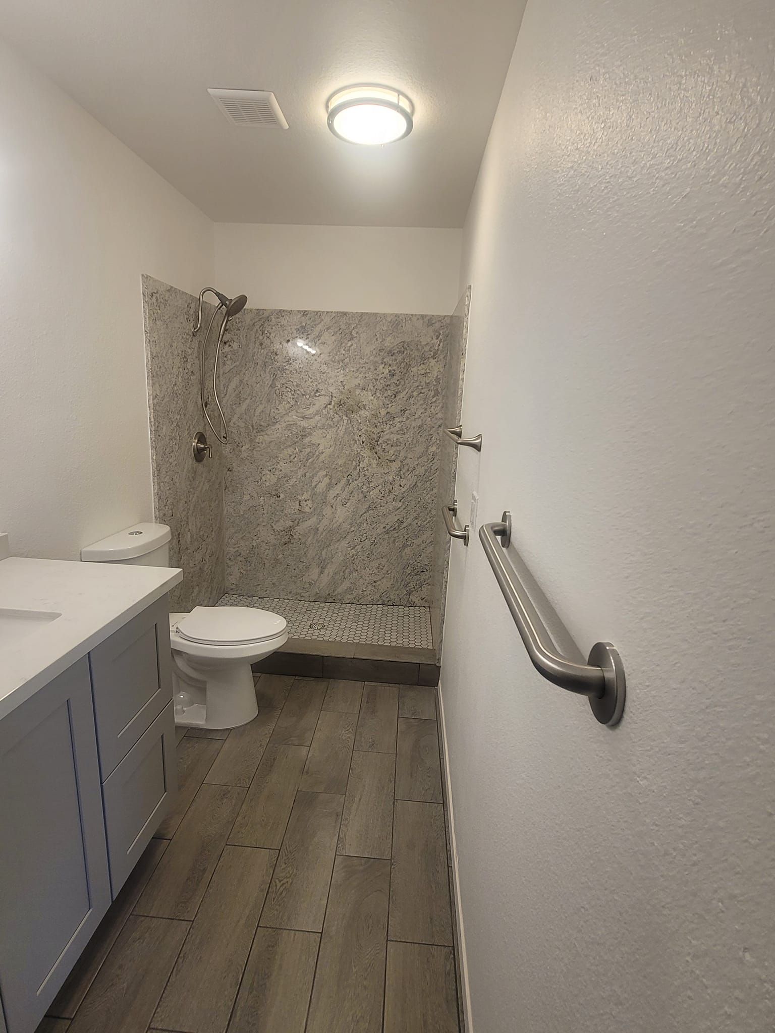 Bathroom with gray wood-look flooring, a white vanity, and a shower with a speckled tile backdrop. A grab bar is mounted on the wall.