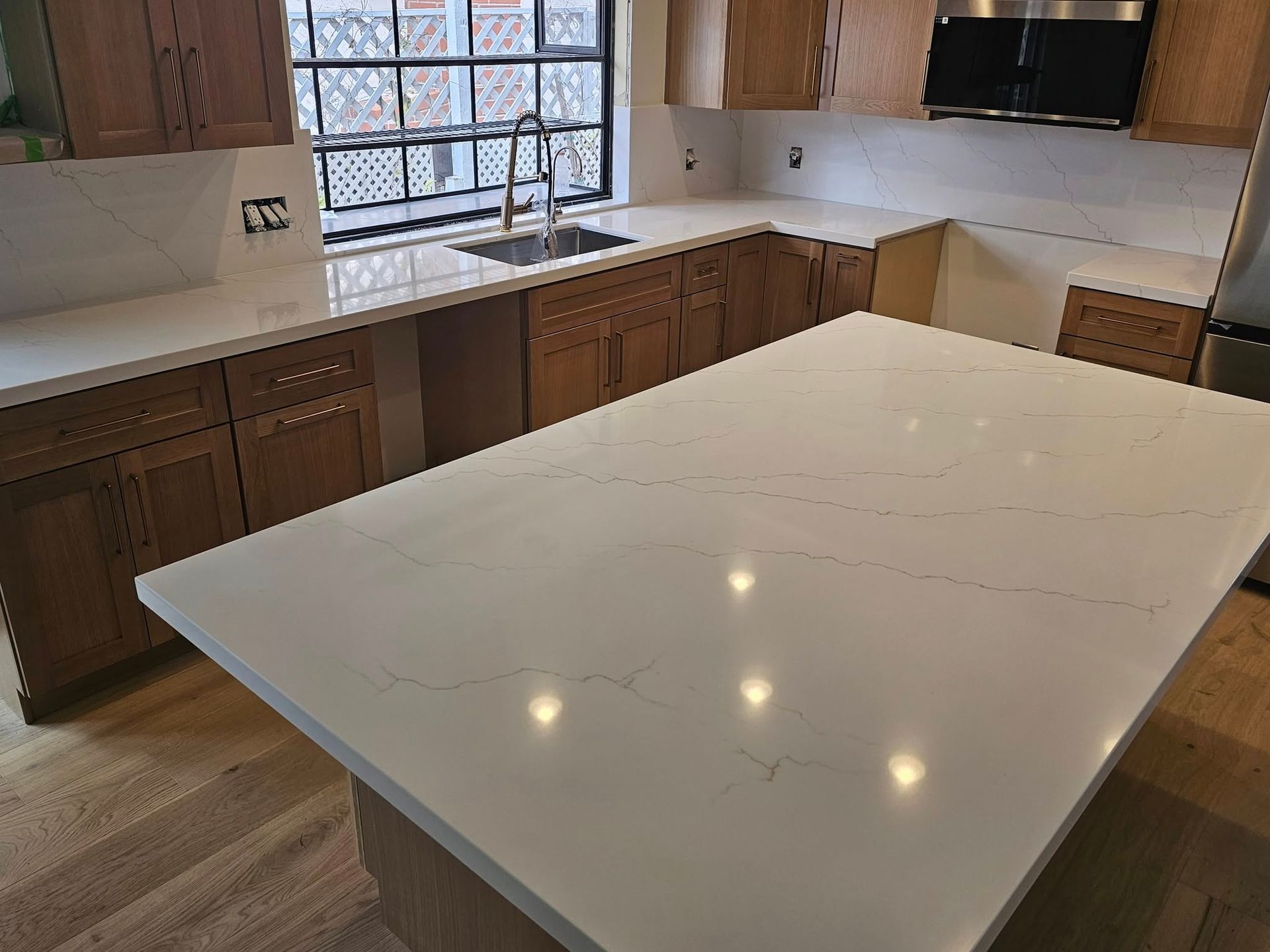 A bright, modern kitchen with light-colored countertops and wooden cabinets. A large island is in the center, and a window is visible.