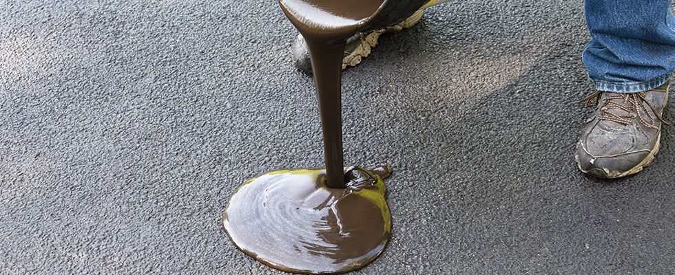 Person pouring dark liquid from a container onto asphalt