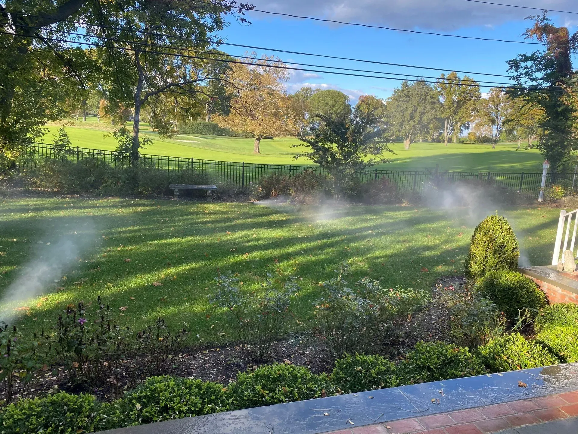 Lawn sprinklers watering a green lawn with a grassy hill and trees in the background under a blue sky.