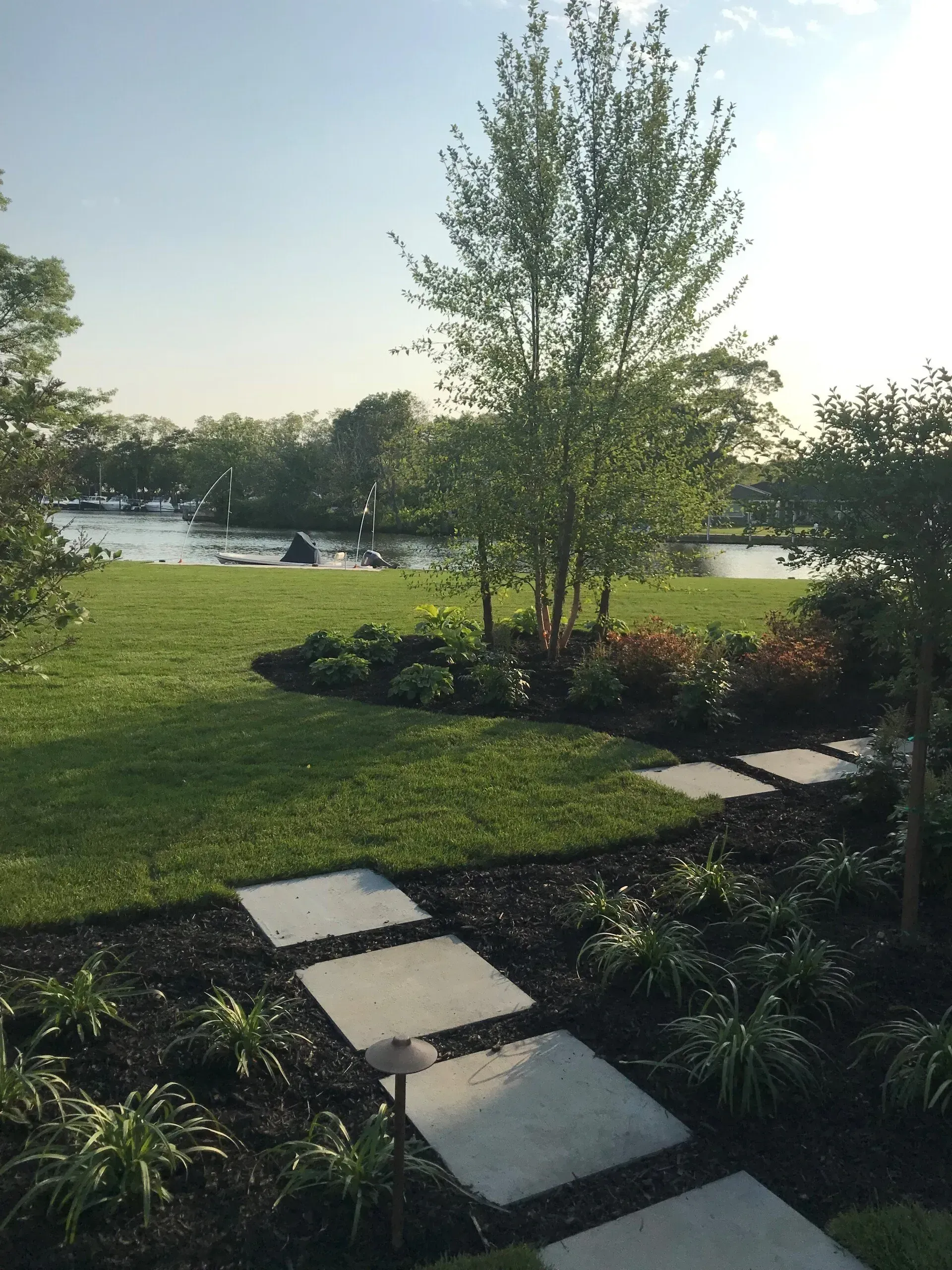 Path of square stones leads through a manicured green lawn to a lake, with a tree and foliage in the foreground.